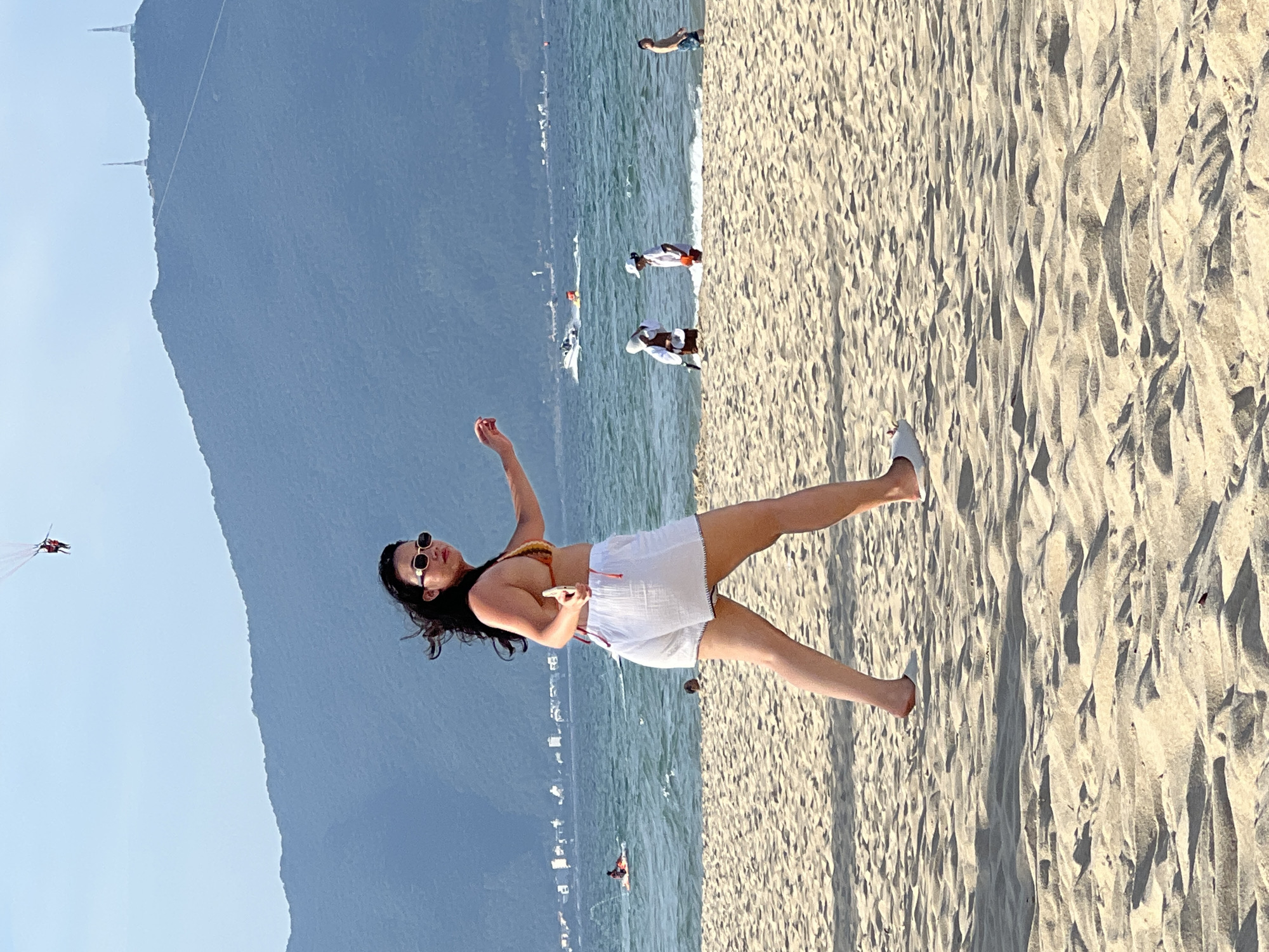A woman with dark, wavy hair and sunglasses, wearing a patterned bikini top and white shorts, walks on a sandy beach in Da Nang, Vietnam. She is in the foreground, her body angled away from the camera and her head turned to look back. Her right arm is extended forward, and she holds a phone in her left hand. The beach is composed of light-colored sand with visible footprints and shadows. In the background, the turquoise ocean stretches to the horizon, with a small boat and several people visible near the water. The distant shoreline is lined with buildings, and a large, blue mountain range forms the backdrop under a clear sky. High in the sky, two people are parasailing.
