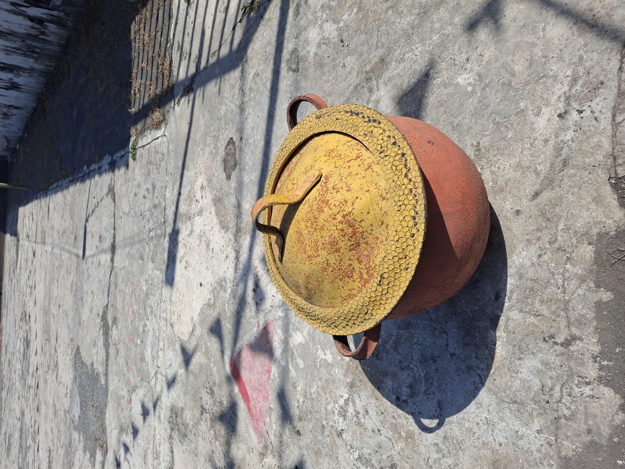 An overhead, medium shot captures a weathered, orange pot with a yellow, textured lid sitting on a cracked concrete surface. The pot has two brown handles on either side of its rim. The lid is a bright yellow, with a distinct honeycomb-like pattern around its edge, and it is visibly peeling, revealing the orange underneath. The concrete is aged and marked with what appears to be faded pink paint and dark stains, suggesting outdoor use. Shadows from an unseen object, possibly a fence or structure, cast a linear pattern across the concrete, indicating it is daytime with sunlight. In the upper left quadrant, a faded red triangular marking is visible on the concrete. The background shows a grey and white wall on the right, also showing signs of age and weathering. The scene is likely located in an outdoor public space in Lumajang, Indonesia, possibly a park or a common area, given the concrete ground and the presence of markings and weathered structures. There are no people or animals visible in the image.