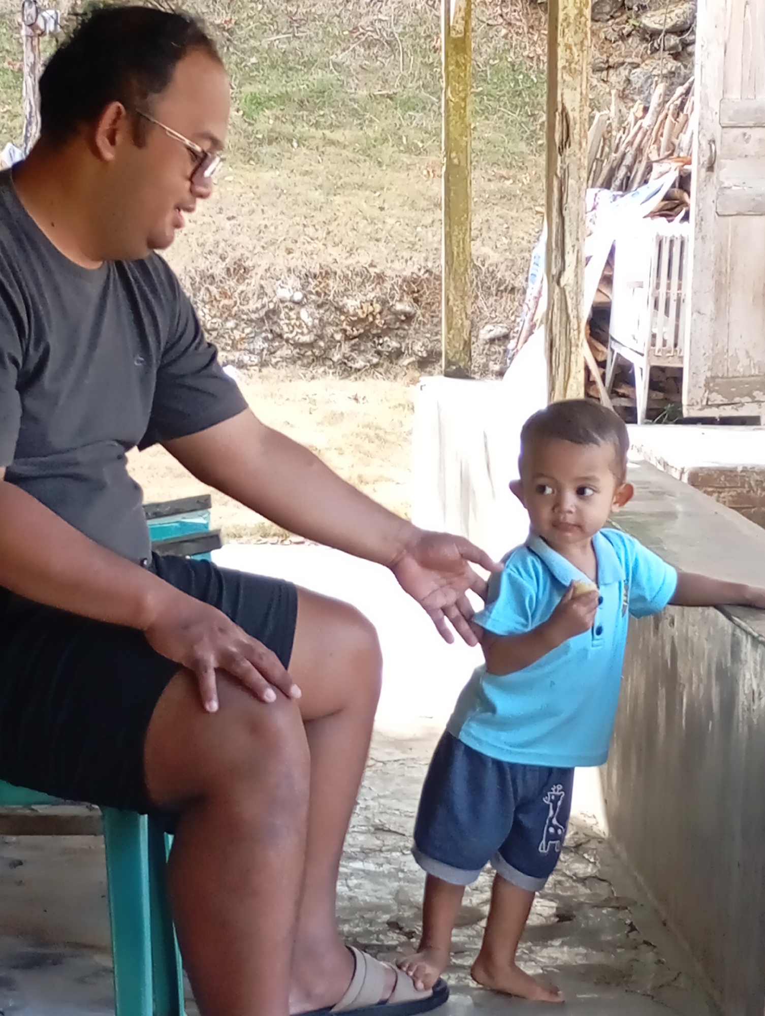In Ketepung, Indonesia, a man wearing a dark t-shirt and black shorts sits on a teal chair, facing left and looking down. His right arm rests on his leg. A young child, wearing a blue collared t-shirt and dark blue shorts with a giraffe graphic, stands to the man's right, leaning against a concrete ledge. The child is holding a small, light-colored object in their right hand, near their mouth, and is looking to their left. The environment appears to be an outdoor area with a backdrop of dry grass and some wooden structures. A wooden pillar is positioned between the man and the child.