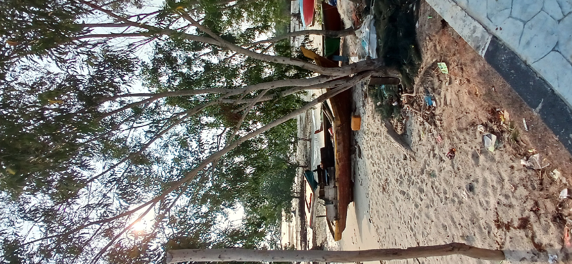 The image depicts a sandy beach area with several boats grounded. In the foreground, a long, wooden boat with a faded red hull and a lighter colored superstructure is prominently displayed, partially covered by a tarp. Nearby, a yellow plastic container is visible. The beach itself is littered with debris, including plastic bags, fishing nets, branches, and various other discarded items. Trees with sparse foliage are scattered across the scene, casting shadows on the sand. In the background, the water is shallow and appears to be receding, revealing a muddy or sandy expanse. More boats, including one with a red hull and another with a green hull, are visible further down the beach. The sky is bright, suggesting daylight, with some sunlight filtering through the trees. The overall scene conveys a sense of a working waterfront or a fishing village that is not meticulously maintained, with evidence of human activity and the passage of time.