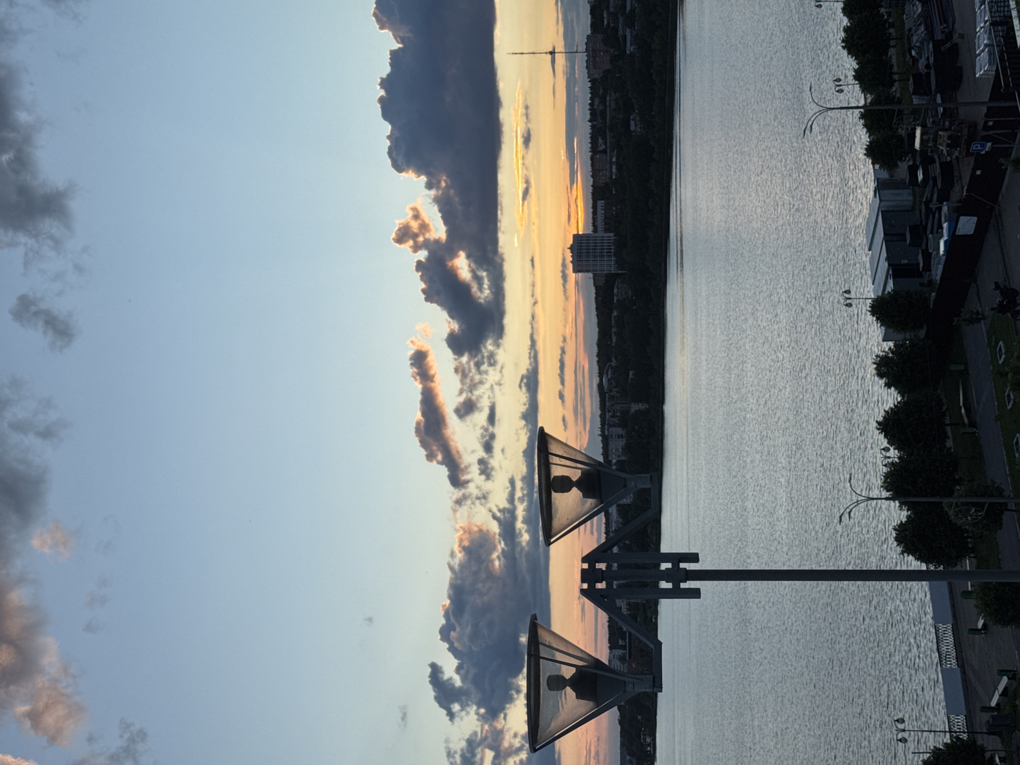This image presents a panoramic view of a waterfront in Izhevsk, Russia, captured during either late afternoon or early evening. The sky, dominating the upper portion, displays a gradient from light blue overhead to warm hues of orange, yellow, and pink near the horizon, where the sun is low. Scattered across the sky are various clouds; some are dark and heavy, while others glow with golden edges, illuminated by the low sun.
In the immediate foreground, two large streetlights with distinctive triangular shades stand silhouetted, framing the scene. Their dark forms contrast sharply with the vibrant sky behind them.
Below the sky and extending into the midground is a broad expanse of water, likely a lake or wide river, its surface textured with ripples that catch and reflect the light.
The distant shoreline across the water features a cityscape with multiple buildings and abundant green vegetation. A prominent, tall, slender structure, possibly a communications tower, rises from the distant horizon. On the nearer embankment, a line of trees runs along the water's edge, interspersed with smaller, multi-branched streetlights. Further back from the water, various low-rise structures, some appearing to be storage units or industrial buildings, and neatly stacked materials are visible.
There are no visible people or specific activities taking place, and no text is identifiable in the image. The overall impression is one of peaceful urban scenery at the golden hour, highlighting the interplay of light, water, and architecture.