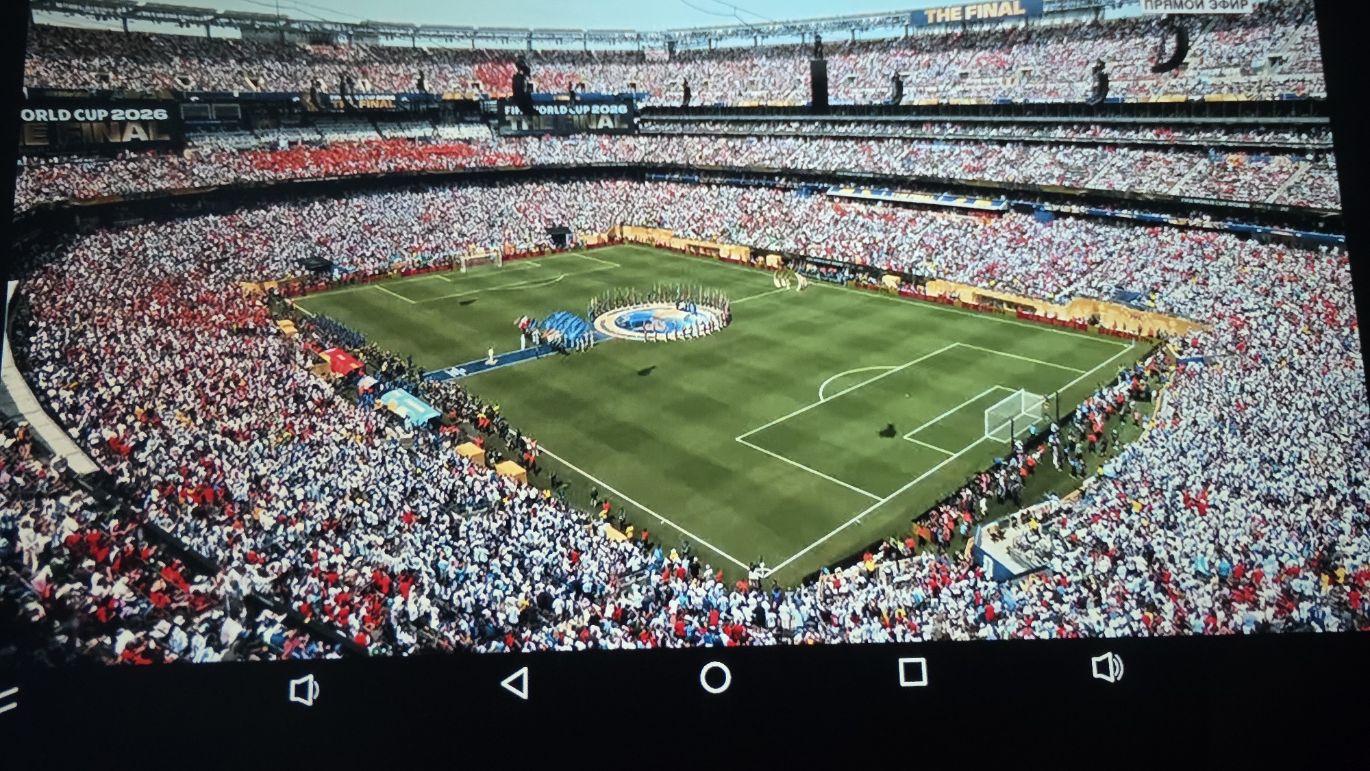 An aerial view shows a packed stadium with a soccer field. The stands are filled with a multitude of spectators wearing primarily red and white clothing, creating a vibrant visual effect. On the field, a circular platform is set up, surrounded by people, possibly for a ceremony or pre-game event. The words "WORLD CUP 2026 THE FINAL" are visible on a banner in the upper portion of the stadium. The scene is captured from a high vantage point, looking down on the action. The image does not provide any specific visual information to determine the location in the city of Kaliningrad, Russia.