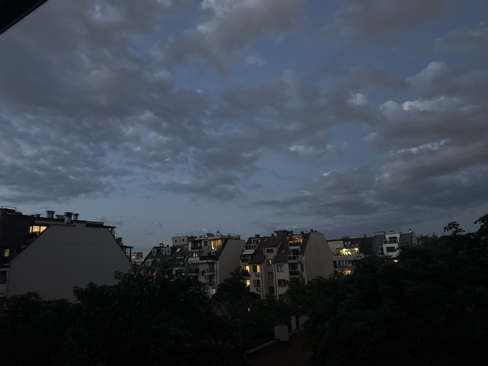 The image shows a cityscape at dusk. Overcast skies dominate the upper portion of the frame, with dark, heavy clouds. Below the clouds, a row of buildings stretches across the horizon. The buildings appear to be residential apartments. Several windows are lit, suggesting it is late in the day. Trees obscure the lower portion of the image. The scene is in Sofia, Bulgaria.