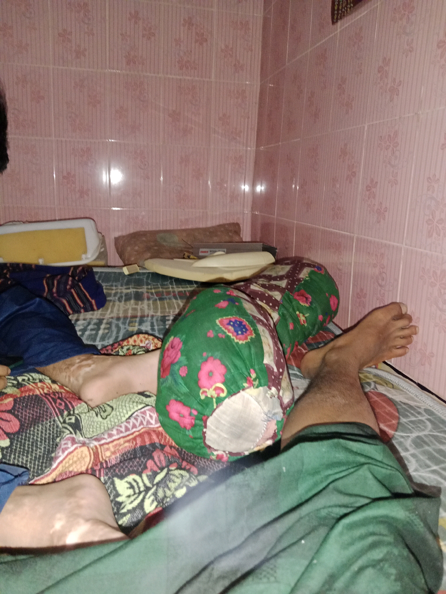 The media file shows a person lying on a bed in a room. The room has pink tiled walls with a floral pattern. The bed is covered with a colorful patterned blanket and a green pillow with a floral design. The person is wearing blue pants and a green garment. One leg and foot are visible. A yellow object, possibly a cushion or mattress topper, is also present on the bed. The lighting suggests it is nighttime or indoors with artificial light. The scene appears to be a domestic setting in Ringinrejo, Indonesia. There is no visible text in the image.
