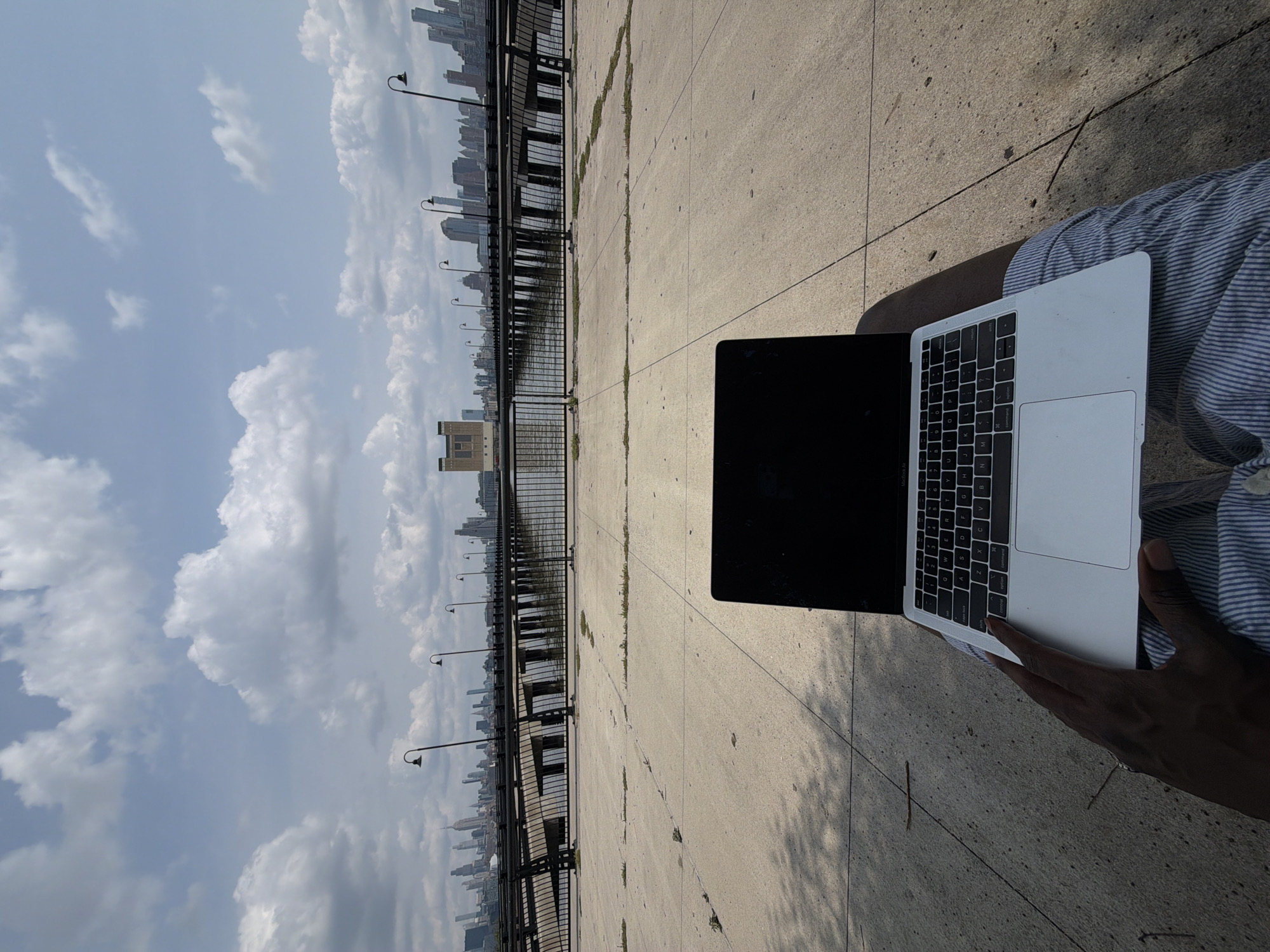 An over-the-shoulder, first-person perspective shot shows a person sitting outdoors with a laptop on their lap. The laptop is silver and appears to be a MacBook. The person's hands are resting on the laptop keyboard and trackpad. In the background, there is a panoramic view of the New York City skyline across the water, with a pier extending into the distance. The pier is lined with lampposts. The foreground consists of a concrete walkway with scattered patches of grass. The sky is partly cloudy with a bright, hazy sun. The overall impression is of someone working remotely or enjoying leisure time with their device in a scenic urban environment. This location is likely Liberty State Park in Jersey City, New Jersey, given the unobstructed view of the Manhattan skyline.