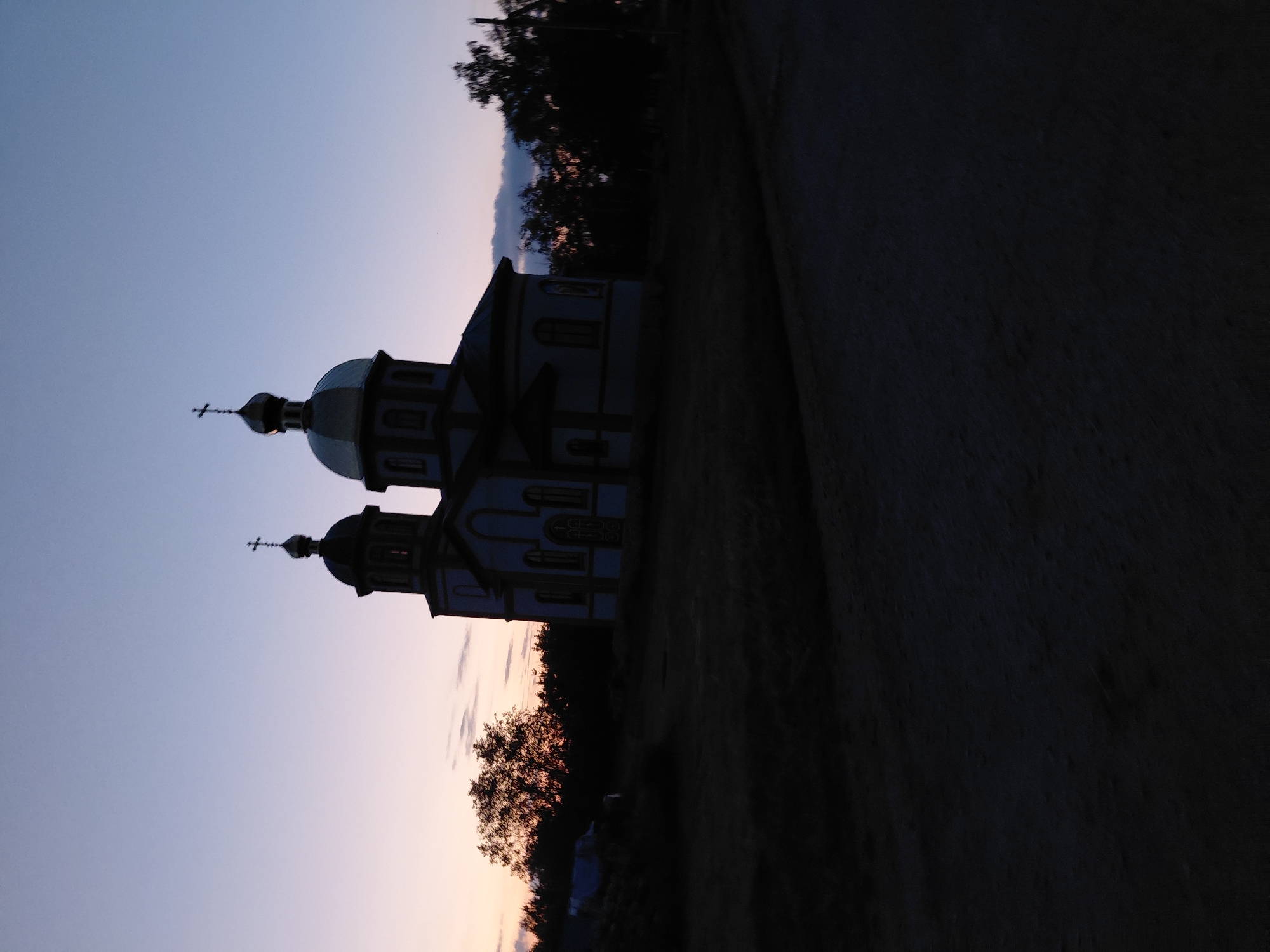 An exterior, medium shot captures a church building silhouetted against a dusky sky in Ustyvytsia, Ukraine. The church, with its distinctive onion dome topped with a cross, stands at the center of the frame. Two domes are visible, one larger and central, and a smaller one to the left. The building appears to be painted a light color, possibly white or pale blue, with darker trim. The sky transitions from a pale pinkish hue near the horizon to a deep blue overhead, with faint clouds visible. Trees and foliage are present to the left and right of the church, appearing dark and indistinct due to the low light. The foreground is dark, suggesting unpaved ground or a dirt road, with some texture indicating unevenness. The overall scene evokes a sense of quiet and twilight.