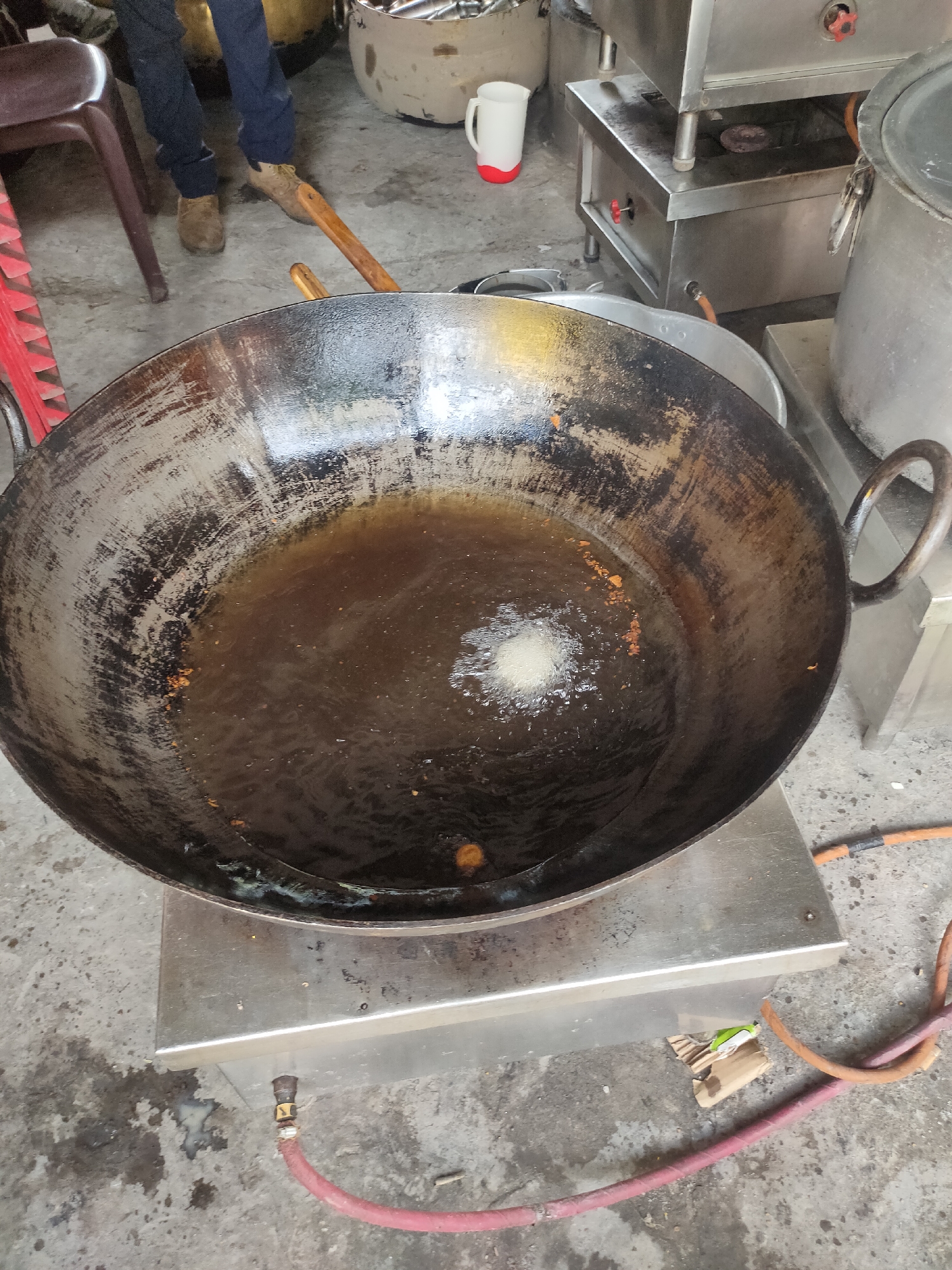 The image captures a scene in Siyal, India, focused on a large, well-used wok filled with cooking oil. The wok rests on a stainless steel stand, which appears to be part of a cooking setup with a gas connection visible via a red hose. A small, round piece of food is actively frying in the center of the wok, causing a noticeable splash of oil. In the background, a person's legs are visible, wearing dark pants and brown shoes, standing near a red chair and a wooden implement. Further back, stainless steel cooking equipment and pots are present, suggesting a commercial kitchen or food preparation area. The floor is concrete and appears somewhat worn and stained.
