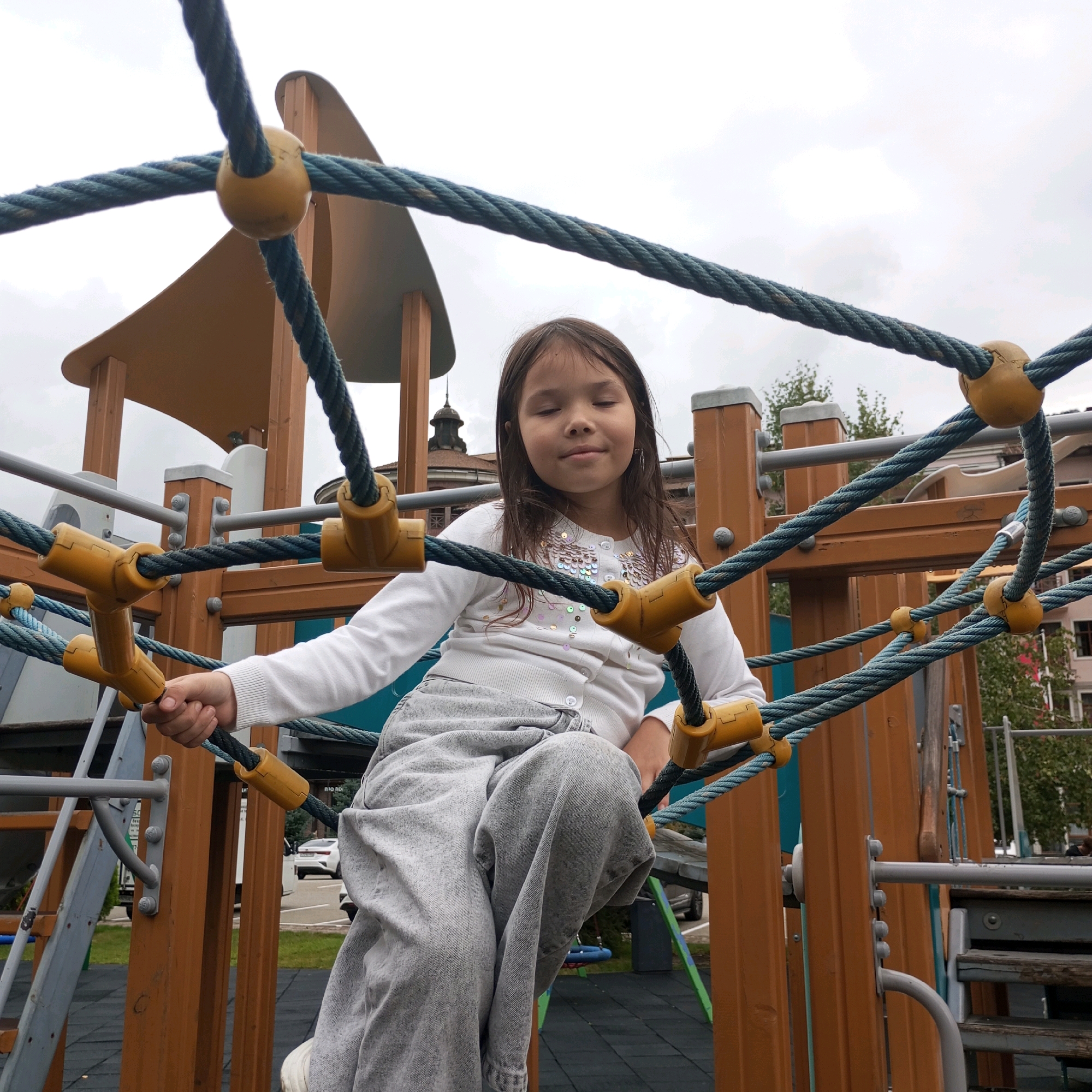 A young girl with long, dark hair is seated on a rope climbing structure at a playground. She is wearing a white cardigan with sequined embellishments and light gray sweatpants. Her eyes are closed, and she has a slight smile on her face. The playground structure is made of wood and thick ropes with yellow connectors. In the background, a building with a distinctive dome and spire is visible, characteristic of architecture found in the resort town of Estosadok, Russia, which is situated in the Krasnodar Krai region. The sky is overcast, suggesting a cloudy day.