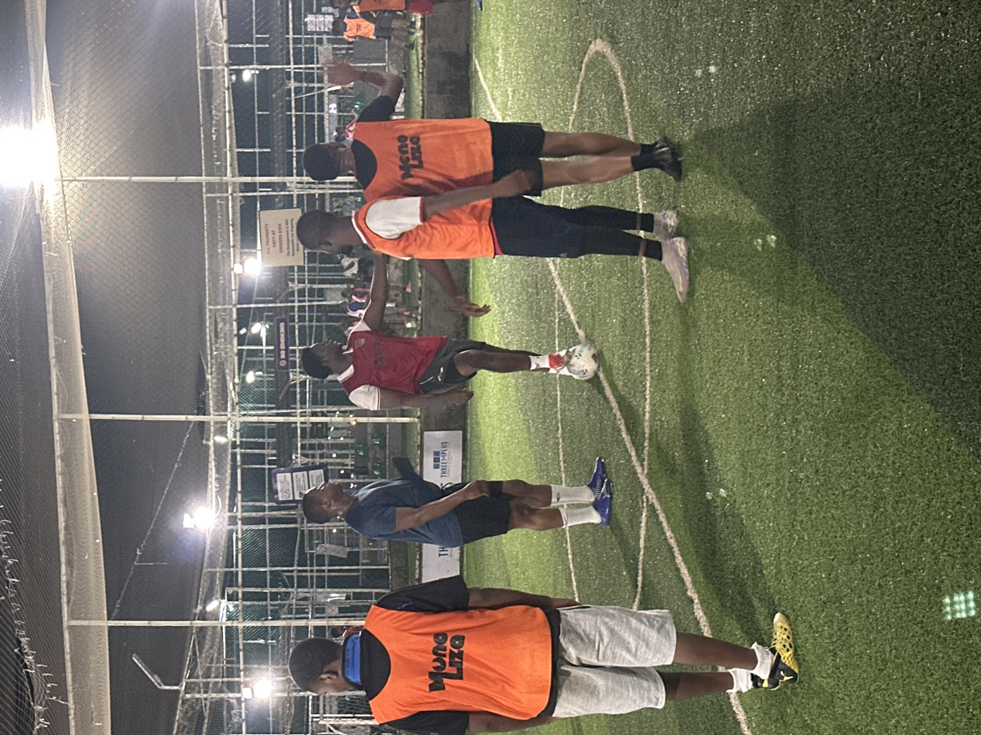 An outdoor, artificial turf soccer field at night, located in Abuja, Nigeria, is the setting for a group of men playing soccer. The field is illuminated by artificial lights, creating a slightly hazy atmosphere. The men are wearing athletic attire, including jerseys, shorts, and soccer cleats. Some are wearing orange bibs over their jerseys, indicating they are part of opposing teams. One man is in the process of kicking a soccer ball, while others are positioned around the field, either watching or preparing to move. The background shows a chain-link fence enclosing the field and some distant structures and lights. The overall scene depicts a casual soccer game taking place after dark.