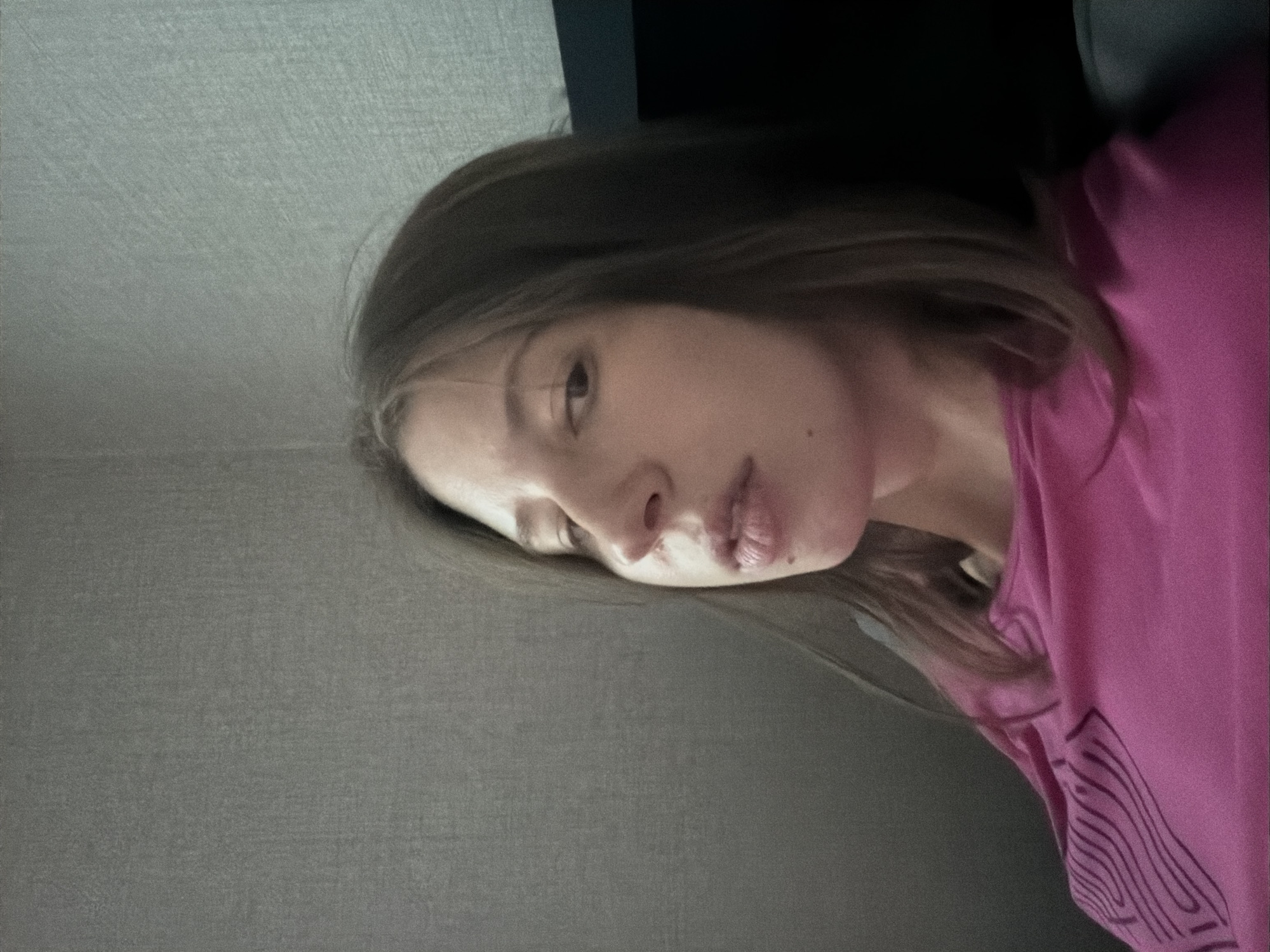 A young woman with brown hair is captured in a medium close-up shot, looking upwards and to the left. She is wearing a bright pink t-shirt with a black abstract pattern on the left side of her chest. The background consists of a textured grey wall, possibly wallpaper, and a dark object to the right, which could be furniture or a wall. The lighting is dim, casting shadows on her face and creating a somewhat grainy texture throughout the image. The setting appears to be indoors. There is no discernible information in the image that places it in Dongducheon-si, South Korea.