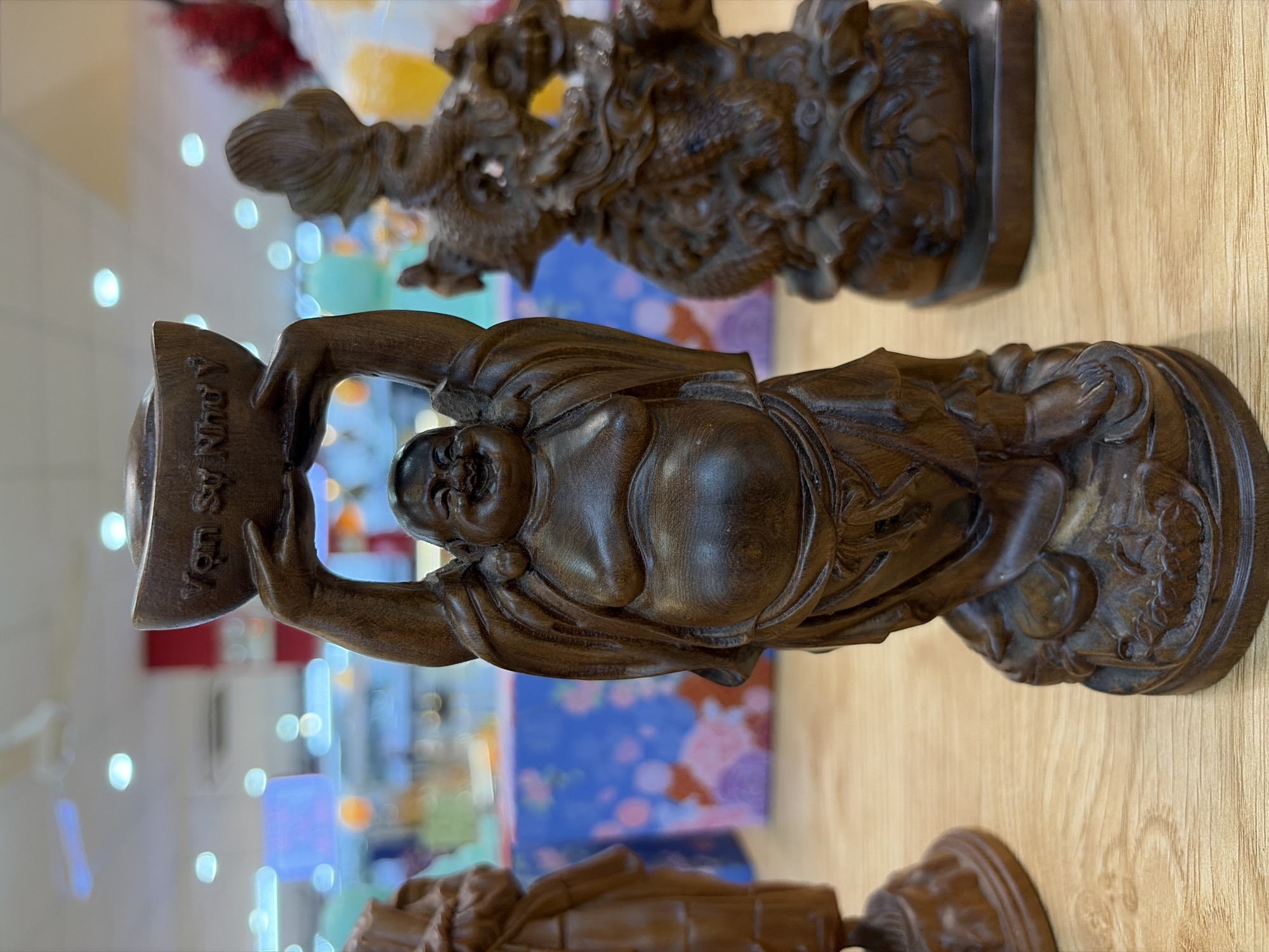 The image shows three wooden statues on a wooden surface. The central and most prominent statue is of a laughing Buddha holding a gold ingot above his head. The inscription "Vạn Sự Như Ý" is visible on the ingot. To the left of the laughing Buddha is another wooden statue, partially visible, appearing to be a standing figure. To the right of the laughing Buddha is a wooden statue of a dragon. In the background, out-of-focus, are store shelves with various items, including what appears to be gift wrapping or packaging with floral patterns in shades of blue and pink. There are also lights and ceiling fans visible in the background. The overall scene suggests a retail environment, possibly a shop selling decorative items or gifts in Đà Nẵng, Vietnam.
