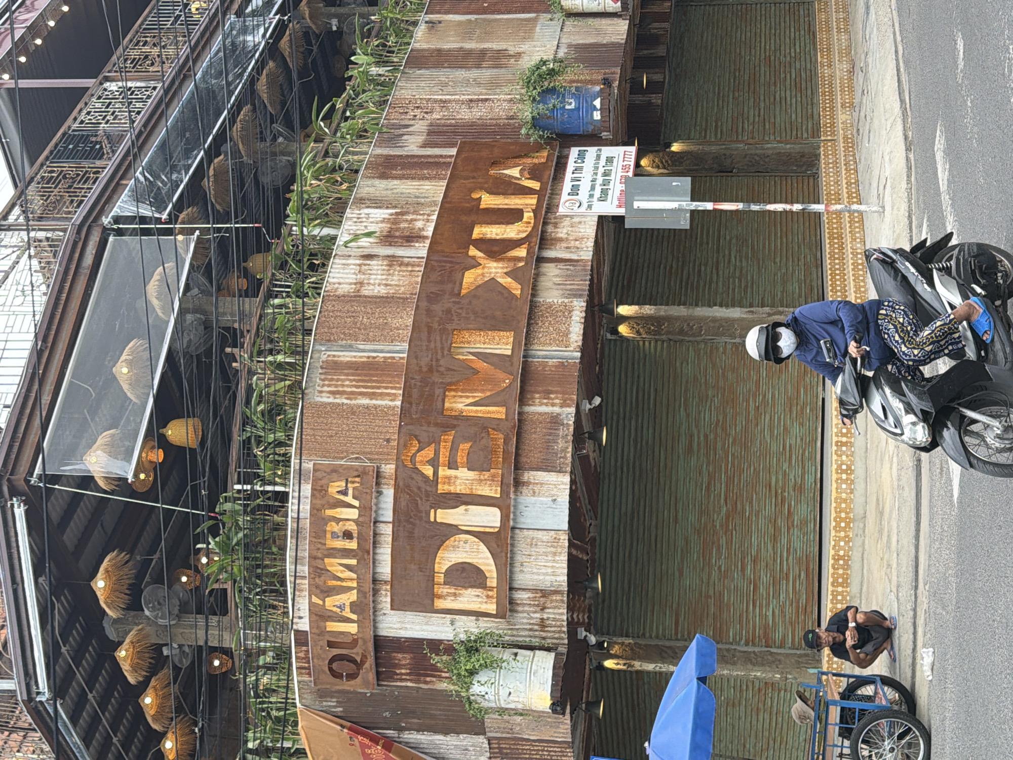 The image shows a street scene in Nha Trang, Vietnam, with a prominent building facade made of corrugated metal and wooden signage. Two signs are clearly visible: "QUAMBIA" on a wooden plank and "DIỄM XƯA" on a larger rusted metal sign. A third sign provides details for "Đơn Vị Thi Công Khang Huy Nha Trang" with a hotline number.
In the foreground, a person on a black motorcycle is parked on the side of the road. The rider is wearing a dark blue jacket, a white helmet, and patterned pants. To the right of the motorcycle, another person is crouched down, appearing to be resting or working, wearing a dark shirt and pants. Behind them, a blue umbrella and a cart with wheels are visible.
The building itself has a rustic and industrial aesthetic, with various levels and balconies adorned with hanging lamps. Greenery is present, with plants growing along the facade. The overall impression is of a commercial establishment, possibly a restaurant or shop, with a unique, weathered appearance. The time of day appears to be daytime, given the lighting. The scene suggests an active street, with the presence of people and vehicles.