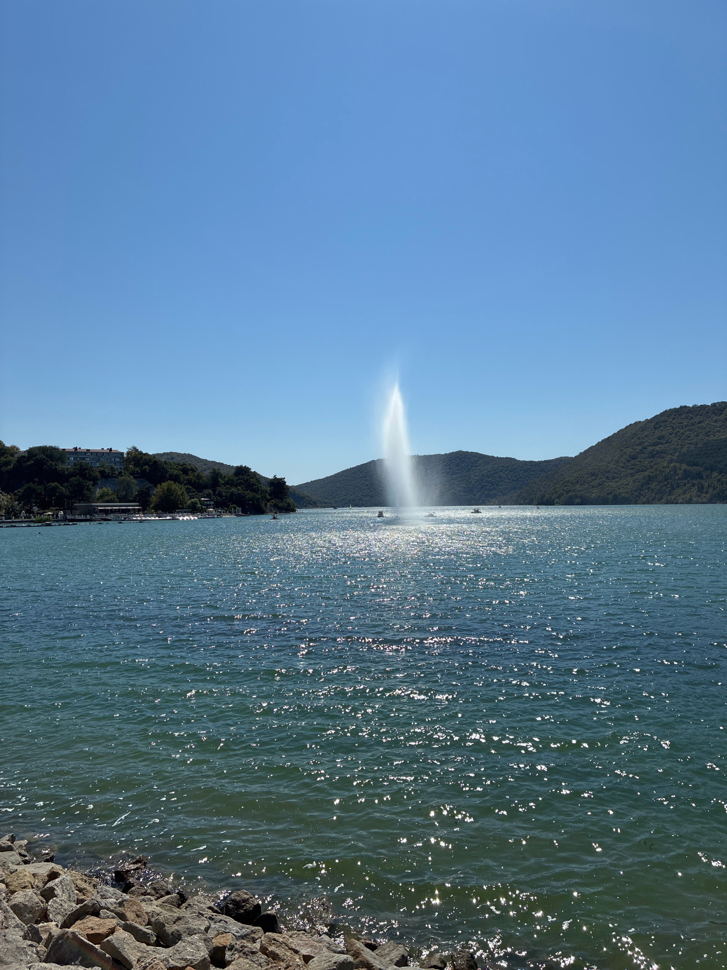 The image depicts a wide, calm body of water, likely the Abrau Lake in Abrau-Dyurso, Russia, on a clear, sunny day. The water reflects the sunlight, creating a sparkling effect. In the center of the lake, a prominent water fountain sprays a tall plume of water into the air. Several small boats are visible in the distance, scattered across the water.
The background features a densely forested, rolling hillside that slopes down towards the water on the right side of the frame. On the left side, the shoreline is lined with trees and some buildings, suggesting a developed area or a resort. The very foreground shows a rocky embankment. The sky is a clear, unblemished blue. There are no people clearly visible in the image.