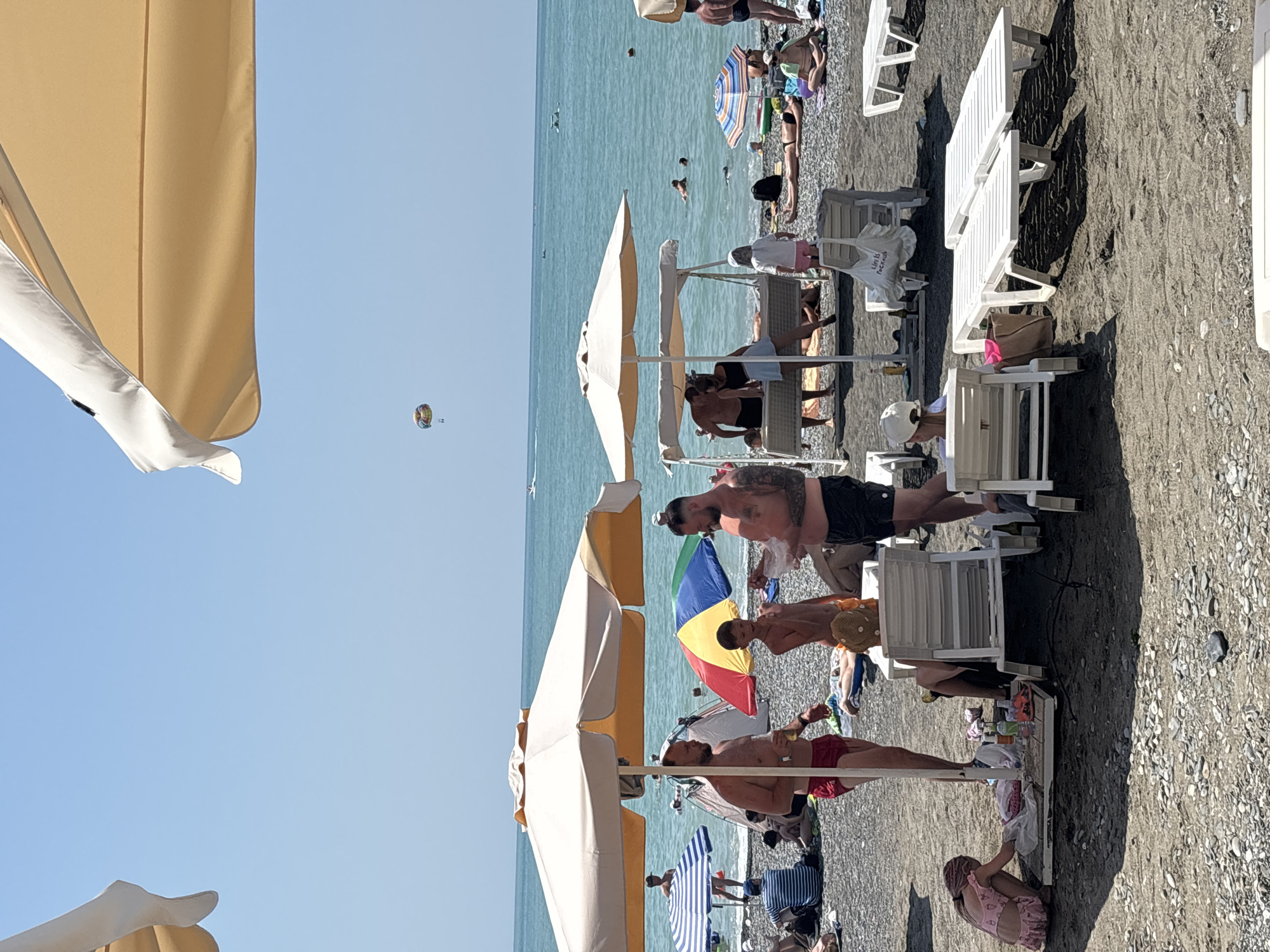 The image depicts a crowded pebble beach on a sunny day. In the foreground, several people are gathered under beach umbrellas and near lounge chairs. A young girl in a pink swimsuit is squatting down, seemingly playing with items on the ground. Two men are standing and talking, one holding a drink. Further back, a swing-like structure has people lounging on it. The sea is visible in the background with several people swimming. A colorful parasail is visible in the sky above the water. The scene appears to be a public beach, likely in Sochi, Russia, given the presence of beachgoers and recreational activities typical of a resort town. The overall impression is one of leisure and summer vacation.