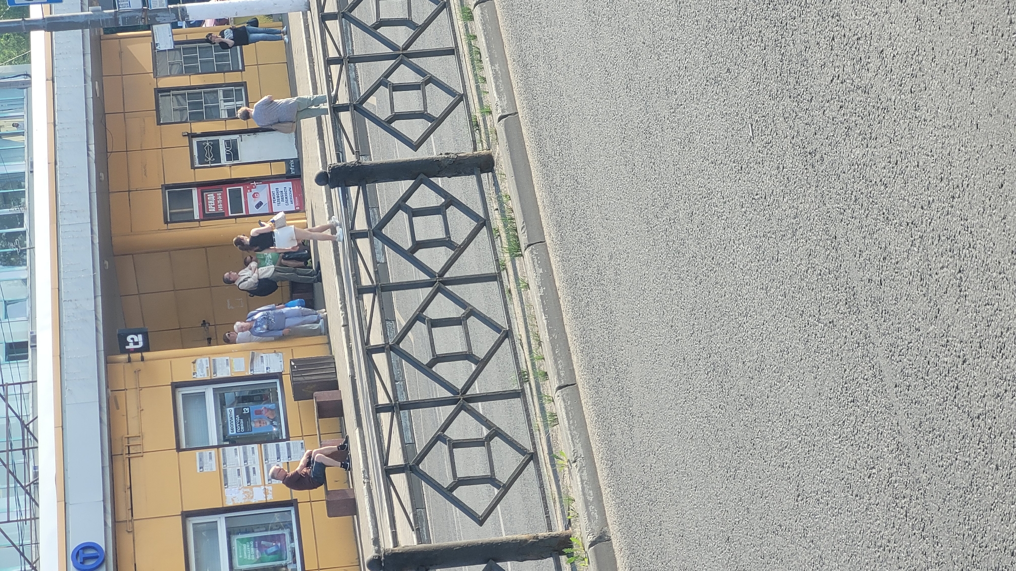 The image depicts a street scene in an unknown city. A yellow building with multiple windows and posters runs along the left side of the frame. Several people are standing or sitting in front of the building, some possibly waiting. A black, decorative metal fence separates the sidewalk from the street. The street, comprising the majority of the right side of the image, is made of gray asphalt. A bus stop sign is visible at the top left corner.