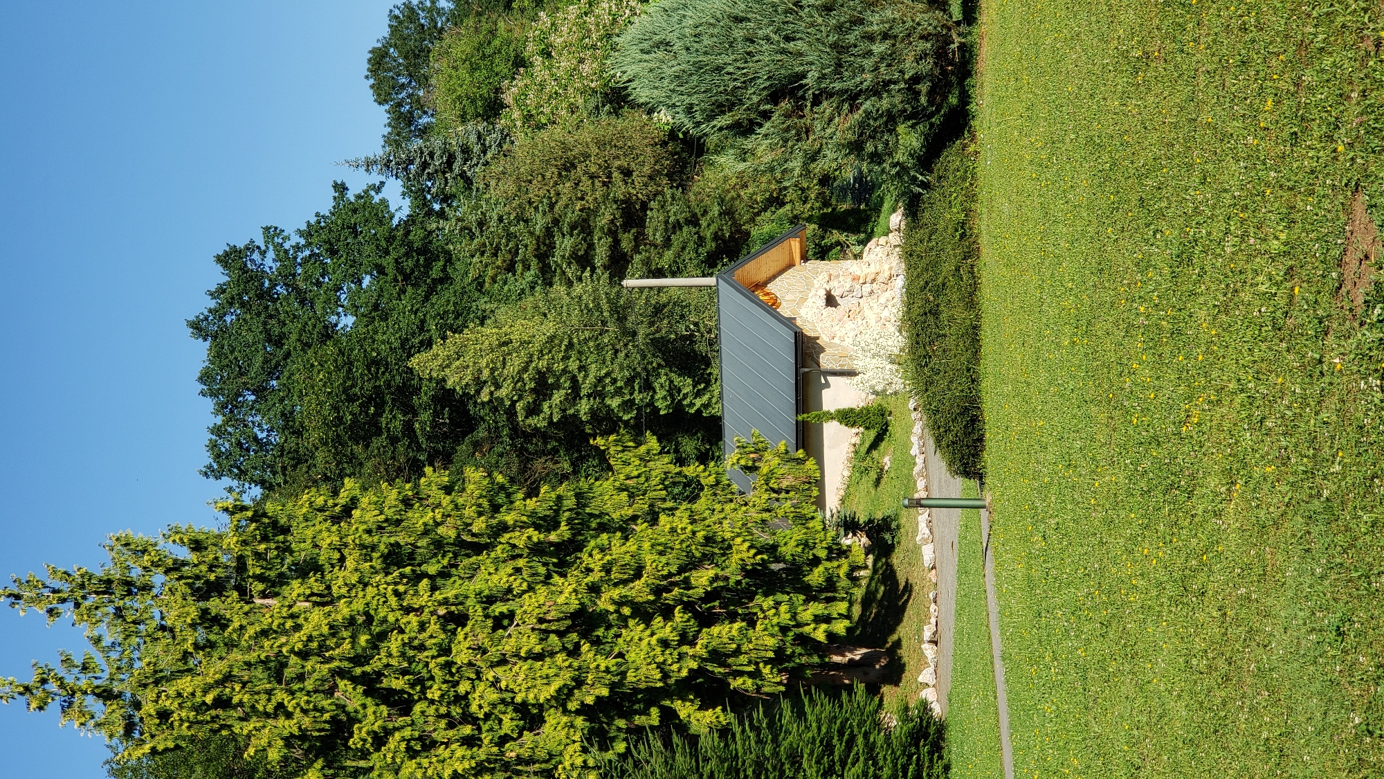 In the city of Prešov, Slovakia, a small stone building with a dark gray metal roof and a chimney is nestled among lush green trees and manicured lawns. The building appears to be an outbuilding or a small shed, constructed with rough-hewn stones and featuring wooden trim around the roofline. In front of the building, a neatly trimmed hedge borders a paved walkway that leads into the scene. The surrounding landscape is a vibrant mix of various trees, including tall conifers with bright green foliage and a dense canopy of mature deciduous trees in the background. A well-maintained lawn, dotted with small yellow and white wildflowers, covers the foreground. The sky is a clear, bright blue, indicating a sunny day. The scene is tranquil and appears to be a park or garden setting within Prešov.