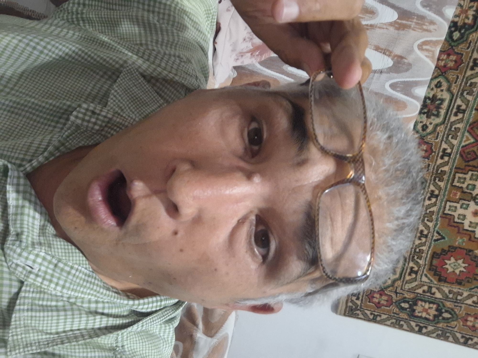A middle-aged man with graying hair and glasses is taking a selfie. He is wearing a green and white checkered shirt. His mouth is open in a surprised expression. The background appears to be an indoor setting with a patterned rug on the wall. There are no visible indicators of his location in Almaty, Kazakhstan.