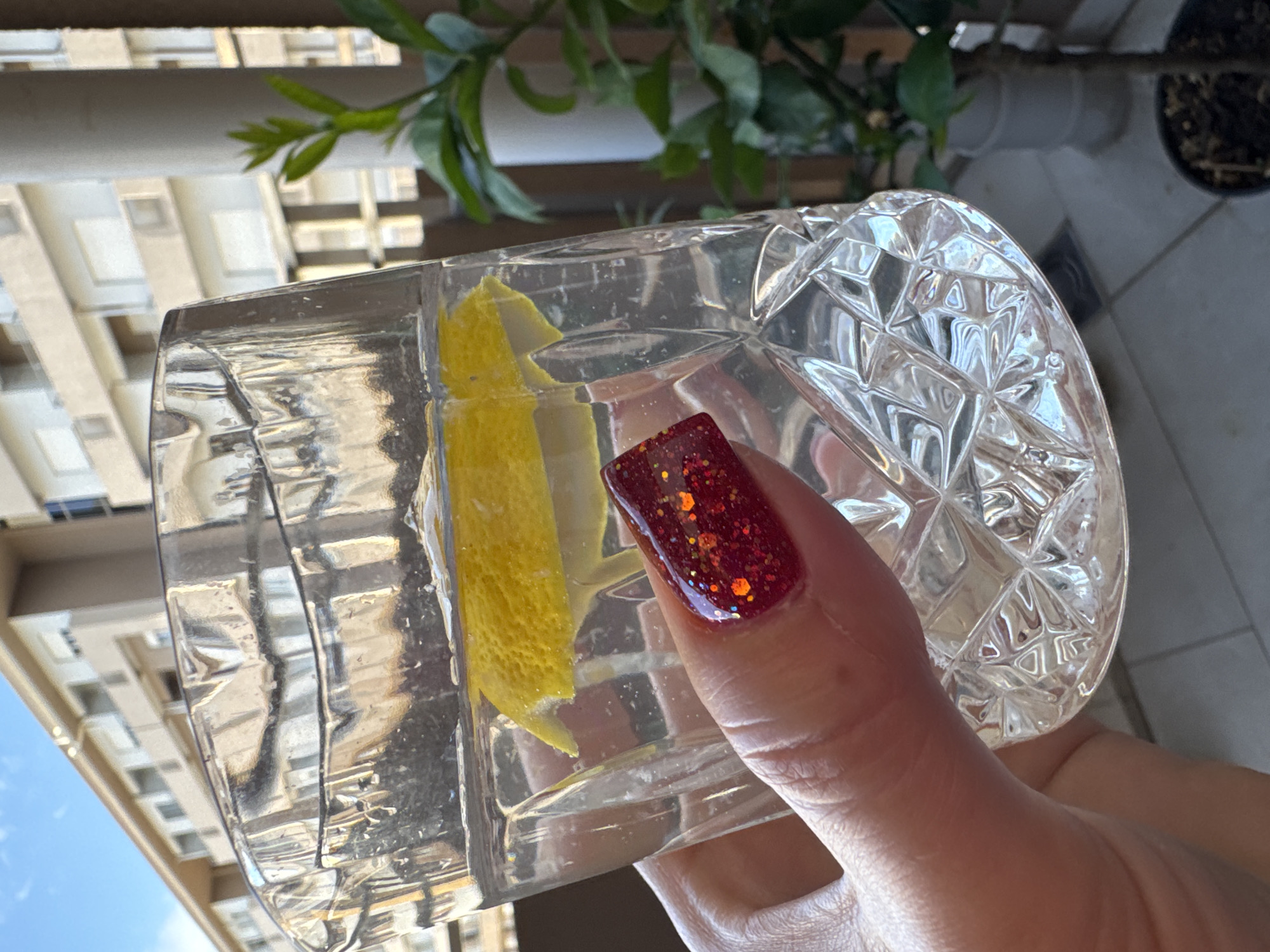 A close-up, eye-level shot shows a hand holding a clear, cut-glass tumbler filled with a clear liquid and a slice of lemon. The glass has a diamond-cut pattern. The hand has a red, glitter-covered fingernail on the index finger, which is pressing against the glass. In the background, out of focus, is a light-colored building with multiple balconies, a blue sky, and a green plant in a pot. The scene appears to be outdoors on a sunny day.