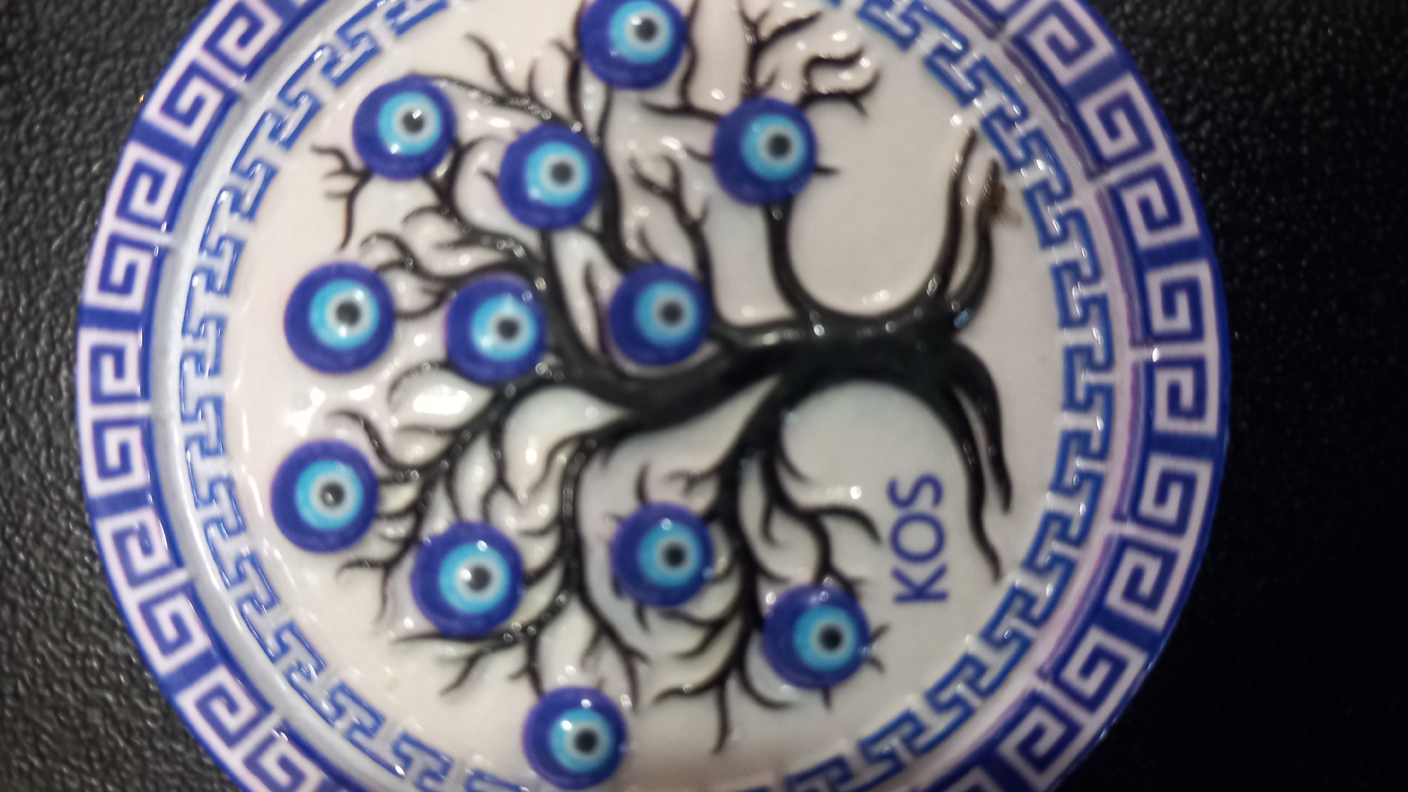 The image shows a round, decorative item with a blue and white Greek key border. The center features a black tree with several blue evil eye charms as its fruit. Below the tree, the word "KOS" is written in blue lettering. This item is likely a souvenir from the city of Kos, Greece, as indicated by the inscription. The background is a dark, textured surface.