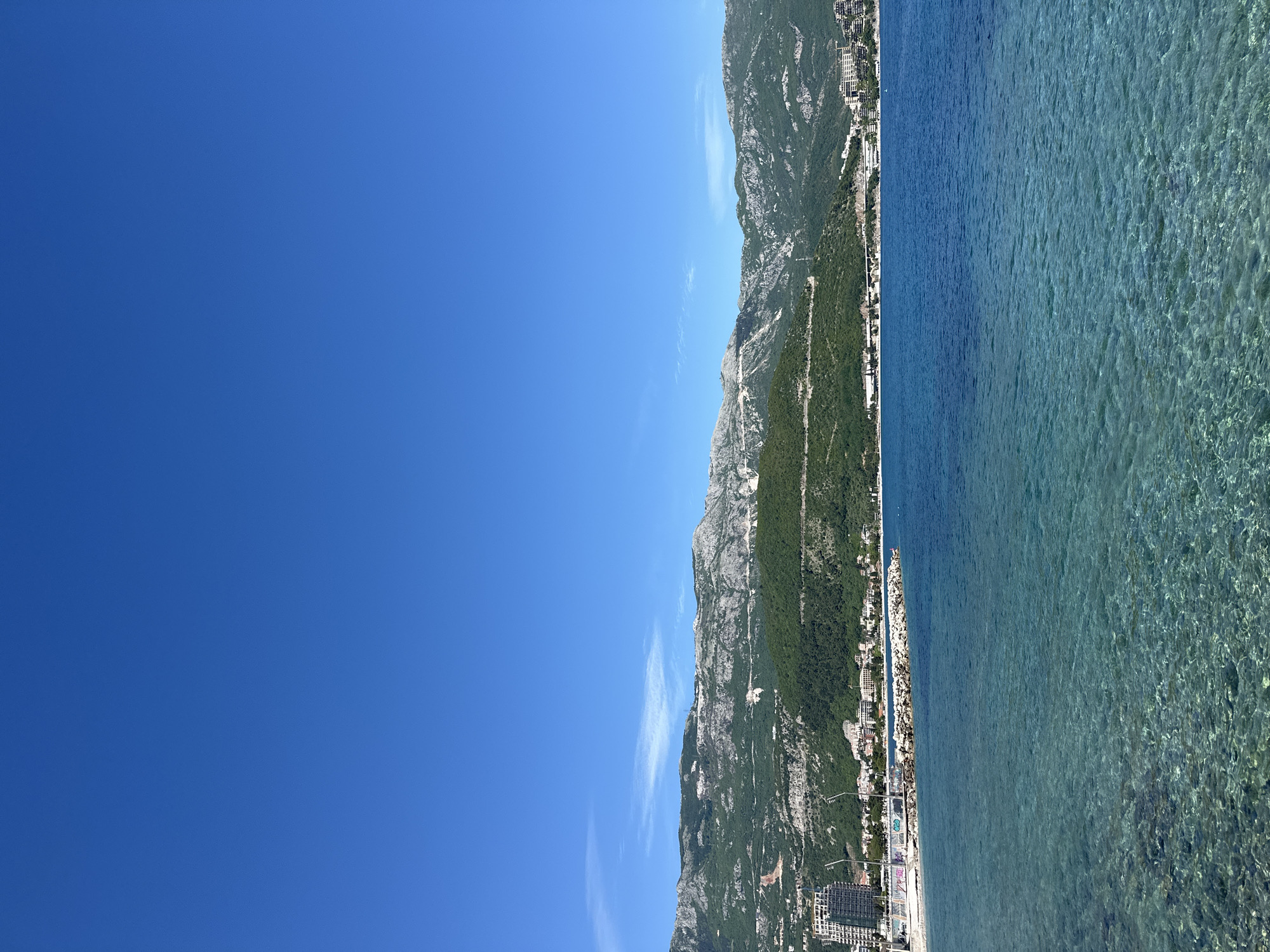 The image shows a scenic view in Budva, Montenegro, featuring a clear blue sky dominating the left half of the frame. A mountain range runs vertically down the center, with lush green vegetation covering its slopes. The right side of the image displays the Adriatic Sea, its surface a mix of turquoise and blue hues, revealing the rocky seabed beneath. Along the shoreline, buildings and infrastructure are visible, suggesting urban development adjacent to the natural landscape. The scene conveys a bright, sunny day at the coast.