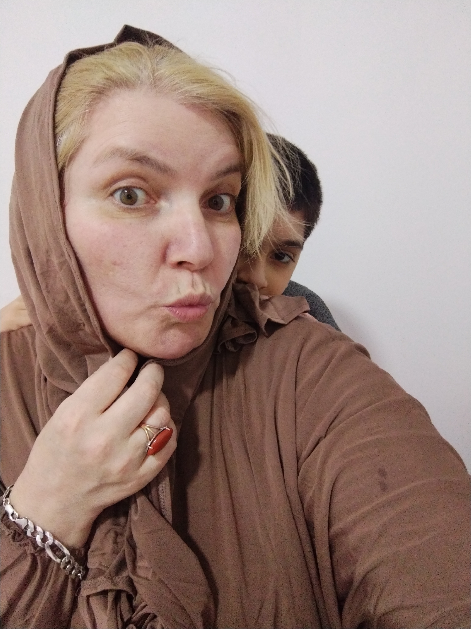 The image is a selfie of two people. A woman with blonde hair and a brown hijab is in the foreground, making a pouting face. She is wearing a brown dress, a silver bracelet, and a ring with a large orange stone. Behind her, a young person with dark hair is peeking over her shoulder. The background is a plain white wall. The scene appears to be indoors. There is no visible indication of the location being in Al Mas'oudi, United Arab Emirates.