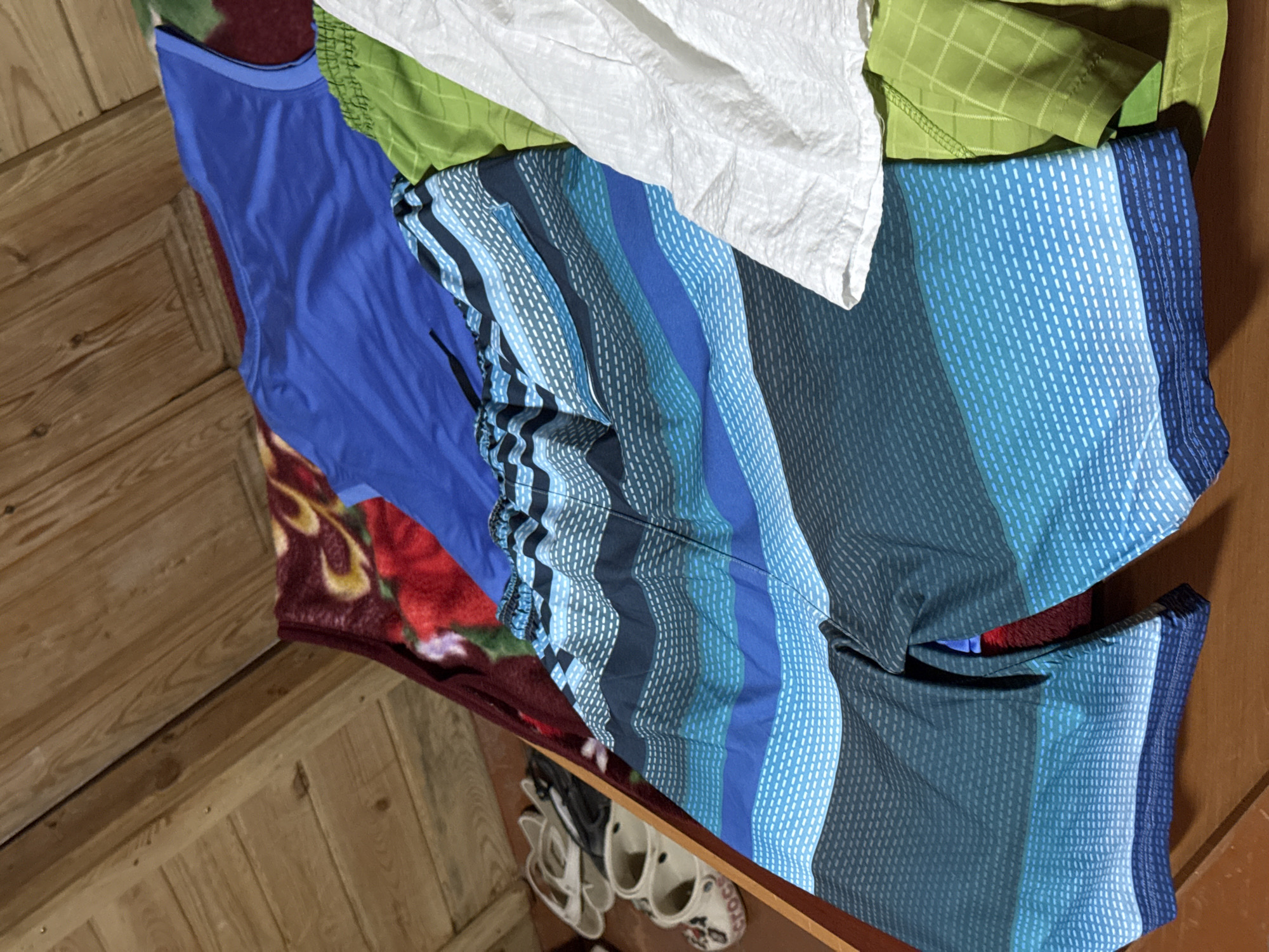 The image shows a pile of clothes, including a blue t-shirt, striped swim shorts, a green t-shirt, and a white crinkled fabric item, laid out on a patterned blanket. To the left, a pair of white Crocs and a pair of white sandals are visible. The background consists of wooden planks, possibly a ceiling or wall, and a wooden surface below the clothes. The scene is indoors, with warm, artificial lighting. There are no people present. The location is identified as Zharbulak, Kazakhstan.