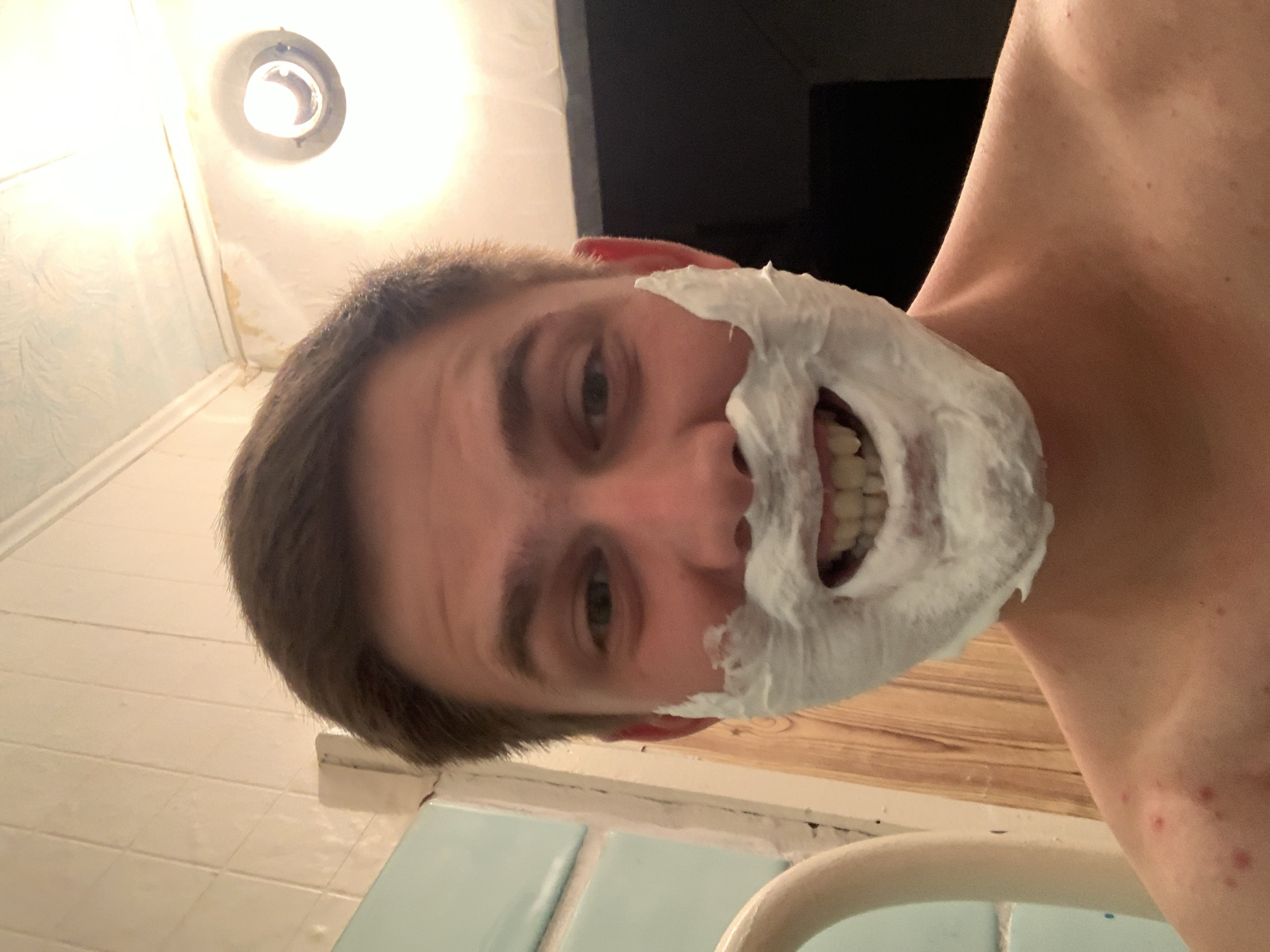 In this close-up, waist-up, point-of-view shot taken indoors, a young Caucasian male with short brown hair smiles broadly, revealing his teeth, while his face is covered in white shaving foam. The foam is applied around his mouth and extends to his cheeks, creating a mustache and beard effect. He is shirtless, and his bare shoulder and upper chest are visible. The background features a tiled wall with a light fixture above, and a wooden surface to the right. A curved, light-colored object, possibly a pipe or railing, is visible on the left side of the frame. The lighting is bright, with a strong light source in the upper right corner casting a warm glow. The scene appears to be a bathroom.