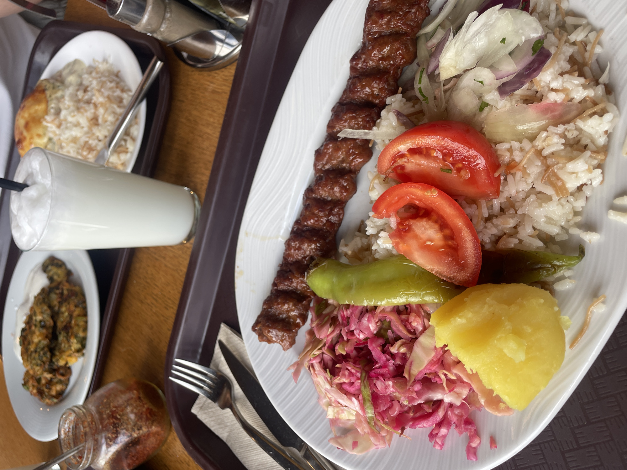 A tabletop view shows multiple dishes served on two brown plastic trays atop a wooden surface within a restaurant. In the immediate foreground, a white oval plate holds a long, segmented skewer of grilled minced meat (kebab), accompanied by white rice pilaf mixed with vermicelli. Side elements on this plate include shredded red cabbage, two slices of red tomato, one whole green chili pepper, a chunk of yellow boiled potato, and sliced red and white onions with green herbs. A silver fork and knife rest on a white paper napkin to the left of this plate.
In the midground, a tall clear glass with a black straw contains a frothy white beverage, consistent with Ayran. To its right, another white plate features white rice pilaf with vermicelli, topped by a golden-brown, baked item, possibly a small casserole or gratin. Behind the foreground plate, a small clear glass jar with a small spoon contains red chili flakes. Further in the background, a third white plate displays three irregular, fried green patties, likely vegetable fritters, topped with a white creamy sauce. A silver-capped salt and pepper shaker set is partially visible on the right.
The culinary style, including the kebab, rice pilaf, Ayran, and specific side dishes, is characteristic of Turkish cuisine. This presentation is consistent with a Gezgin Kebab restaurant, located in Sofia, Bulgaria.