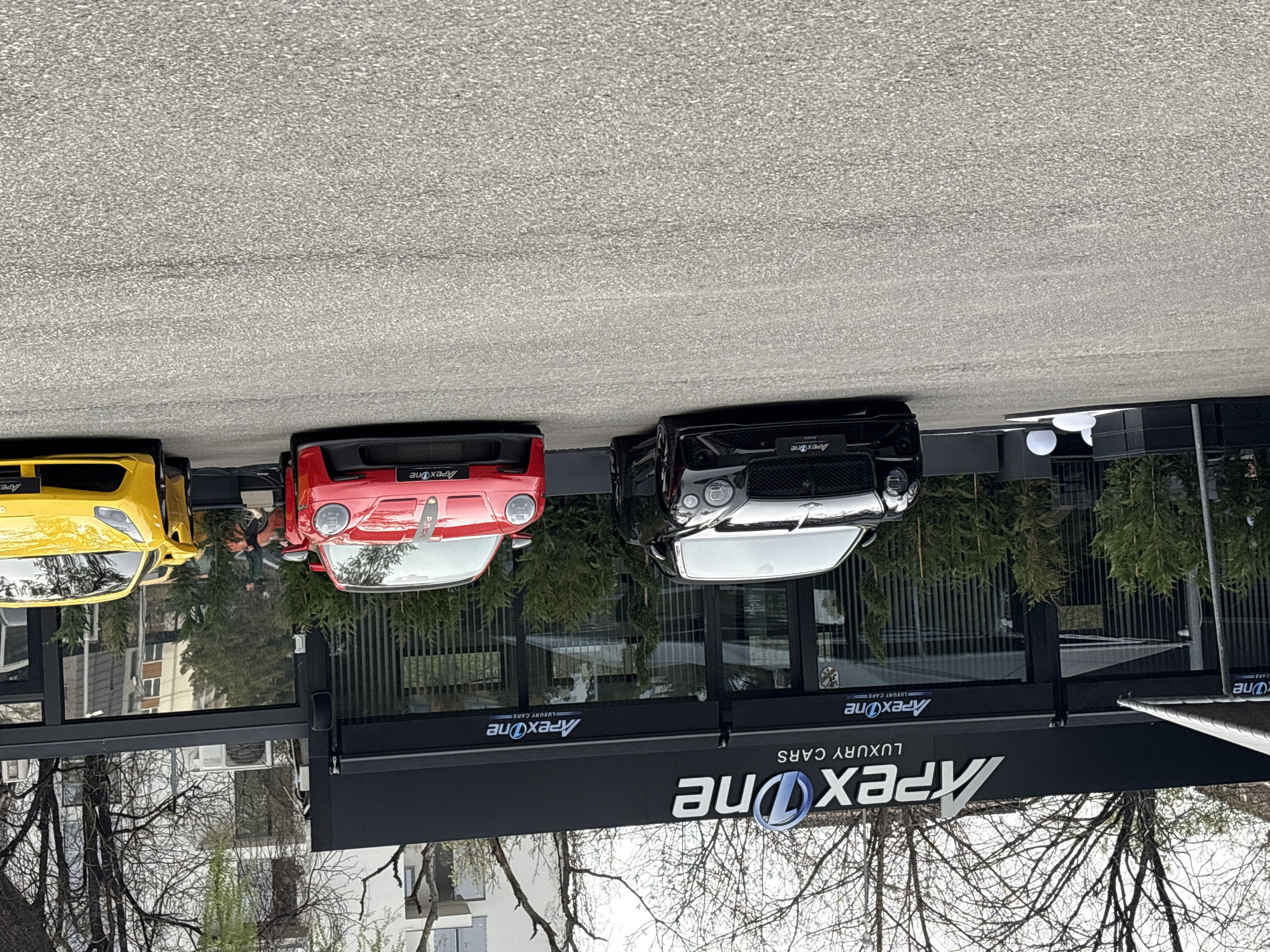 The image shows several luxury cars (yellow, red, black) parked in front of a building with a sign that reads "Apex One Luxury Cars." The building is likely a dealership. The image is taken from a slightly elevated perspective, looking down onto the cars and building. In the background, there is a view of trees and other buildings, suggesting an urban setting. The location is Sofia, Bulgaria, based on the context provided.