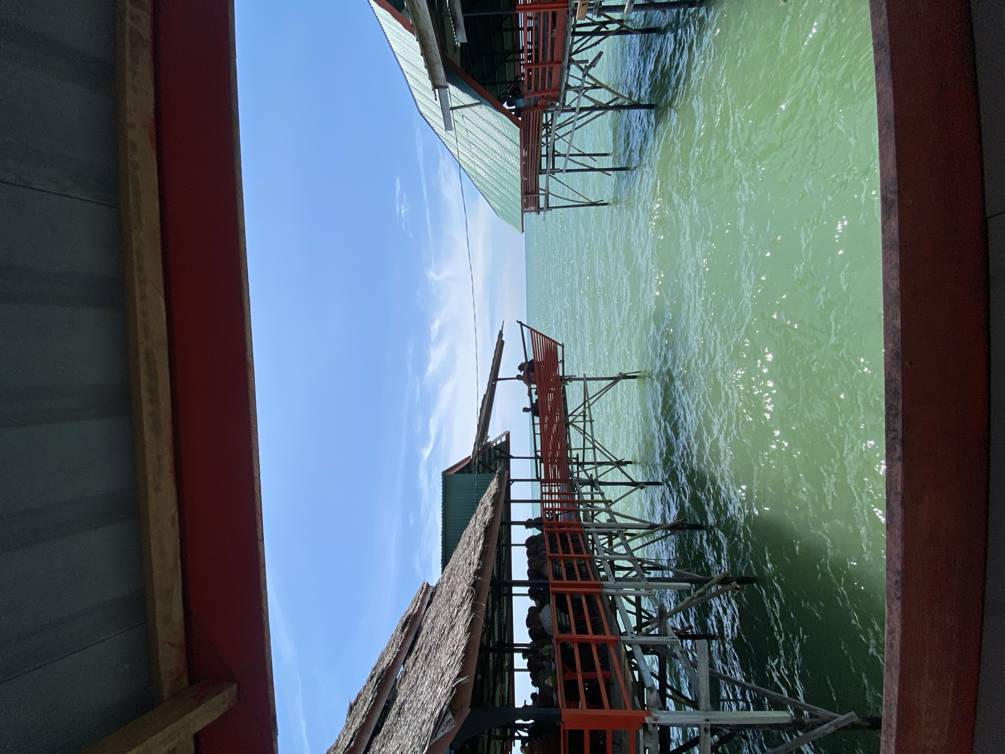 This image captures a scene at a waterfront restaurant in Lancok, Indonesia. The perspective is from within a covered area, looking out towards two open-air dining structures built on stilts over the ocean. The structures have thatched and metal roofs, with vibrant orange railings and wooden support beams. The water is a shimmering greenish hue, with sunlight glinting off its surface. Several people are visible seated at tables within the dining areas, suggesting a bustling atmosphere. The sky is a clear blue with scattered white clouds. The overall impression is of a casual, seaside dining experience.