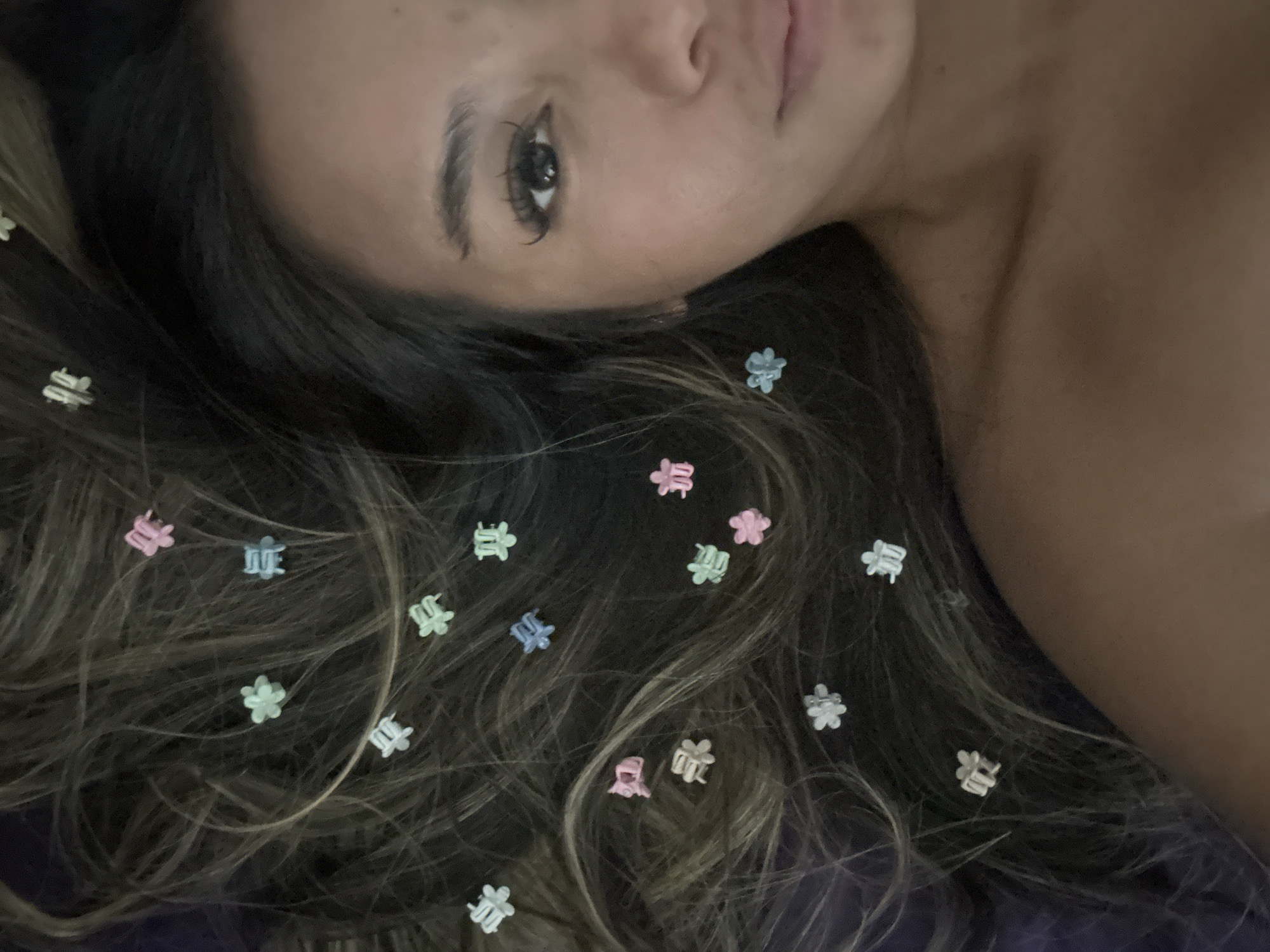 A woman lies on a purple surface with her head turned towards the camera. Her long, dark hair is spread out around her and adorned with numerous small, colorful hair clips shaped like flowers. The background is out of focus. It is not possible to confirm the location in the city of North Jakarta, Indonesia, from the provided image.