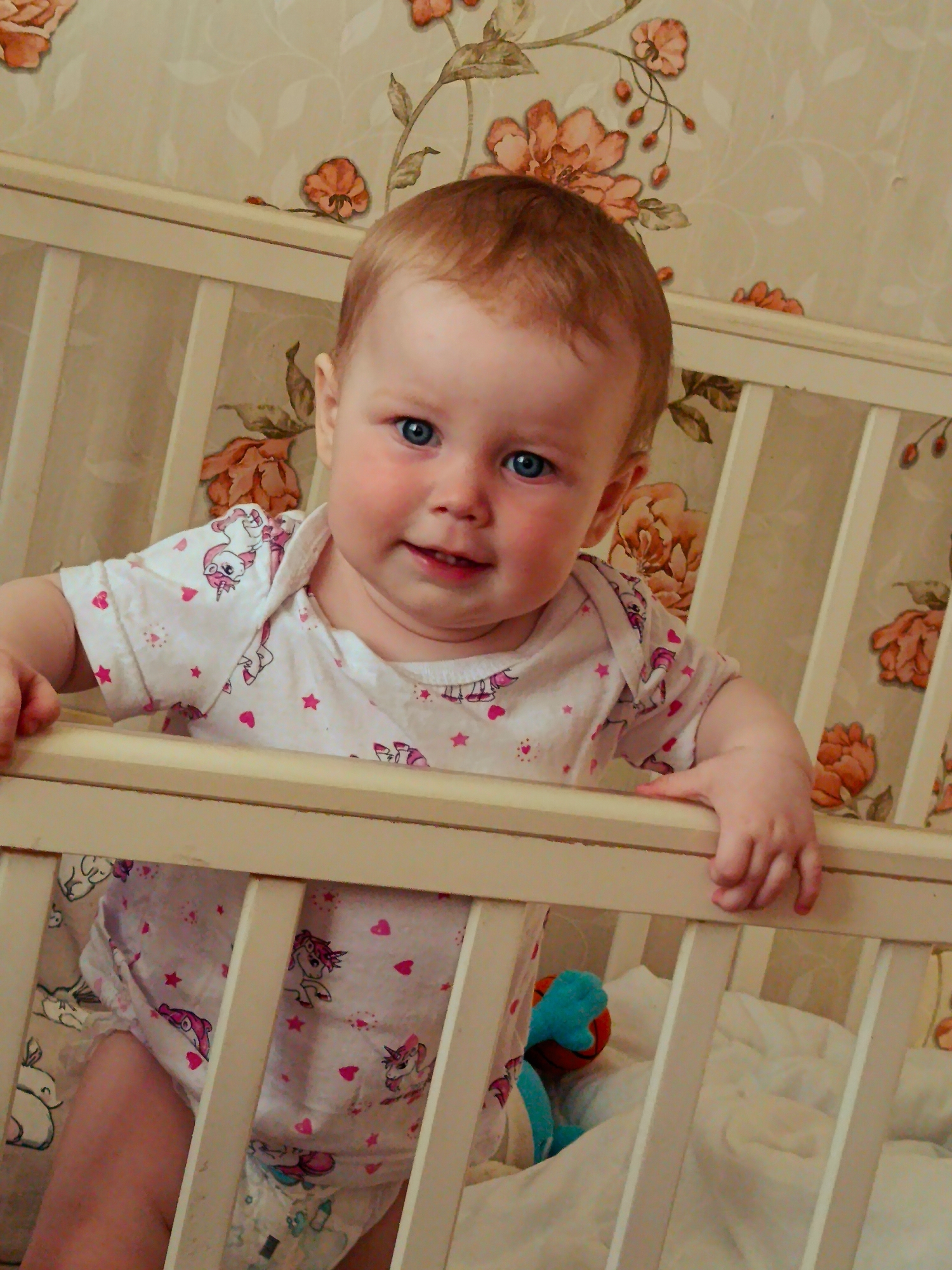 A baby is standing in a crib. The baby has fair skin, blue eyes, and reddish-blonde hair. They are wearing a white onesie with a unicorn and heart pattern. The crib is made of wood and painted white. There is a floral wallpaper behind the crib. The scene is indoors. Location Фоки, Russia is not evident.