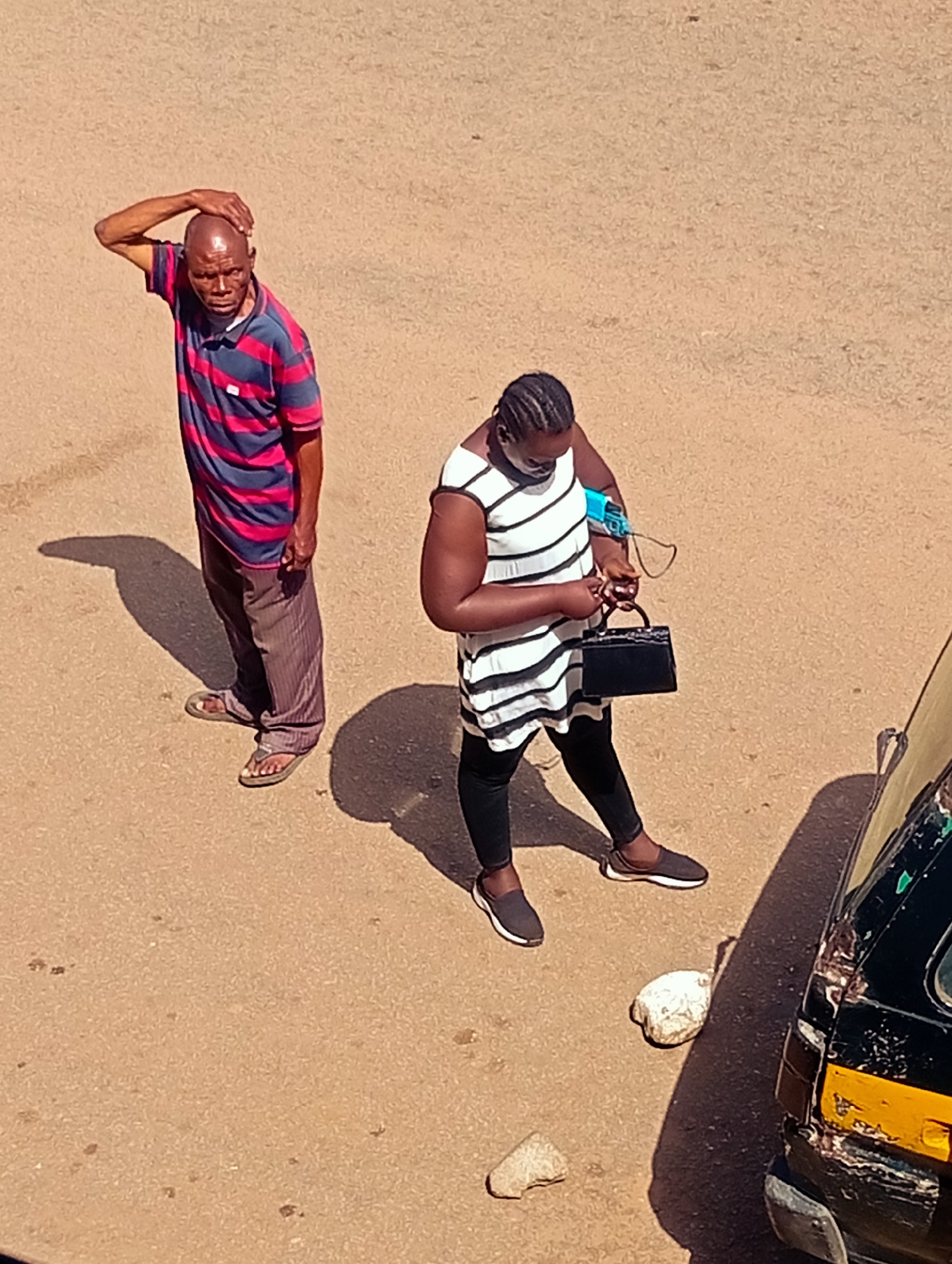 A man and a woman are standing on a dusty, tan-colored ground, possibly a road or open area. The man, on the left, is bald and older, wearing a red and blue striped polo shirt and brown striped trousers. He has his right hand on the back of his head, appearing to be in thought or perhaps feeling stressed. He is wearing sandals. The woman, on the right, is younger and wearing a black and white striped sleeveless dress over black leggings. She is holding a small black handbag and appears to be looking down at it, possibly checking its contents or adjusting something. She has braided hair and is wearing dark shoes. To the right of the woman, a portion of a vehicle, likely a taxi, is visible. It is black with a yellow stripe along the back and shows signs of wear and tear. The scene suggests a daytime setting, indicated by the strong shadows cast on the ground, implying a sunny day. The location appears to be Bukuru, Nigeria, and the overall atmosphere is one of everyday life on a street or marketplace. There is no visible text in the image.