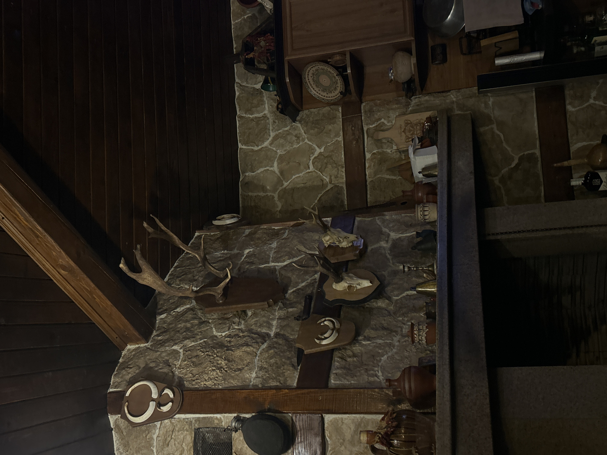 The image captures an interior wall adorned with hunting trophies and rustic decor, likely within a building in Mirovyane, Bulgaria. The wall is constructed from irregular stone pieces, giving it a textured appearance. Mounted on the wall are several sets of animal antlers, along with plaques featuring animal skulls or tusks. These items suggest a hunting theme. Other decorative objects include pottery, a large pan, and wooden accents. The area is dimly lit, with dark wooden paneling visible in the background on one side of the wall, contrasted by another side that has the rustic wall with hunting artifacts.