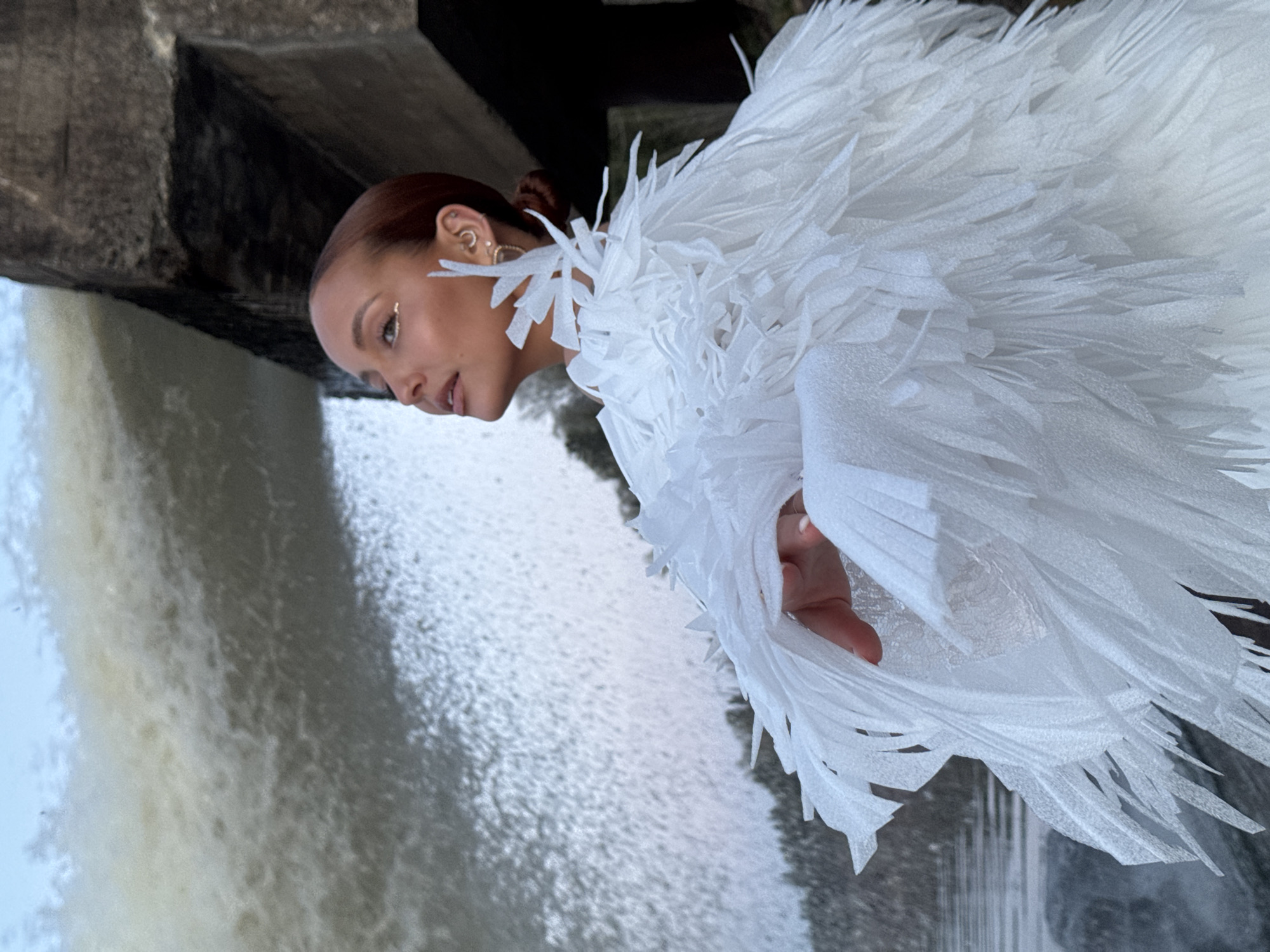 The image shows a woman with reddish-brown hair pulled back into a bun, wearing a striking white garment resembling feathers. She stands in front of a waterfall cascading from a concrete structure. The location is Noviy, Russia. She wears makeup with what appears to be a gold tear drop under her right eye. The angle is slightly tilted. She is looking at the waterfall.