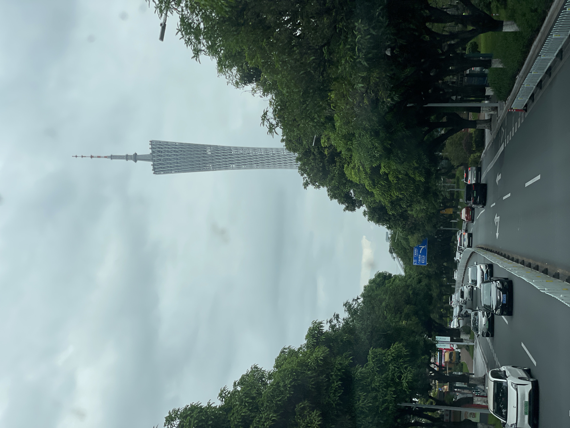 The image depicts a scene on a road in Yuexiu District, China. The Canton Tower (also known as Guangzhou TV Tower) is visible in the distance. Cars are driving on the road, which is lined with trees on either side. The sky is overcast.