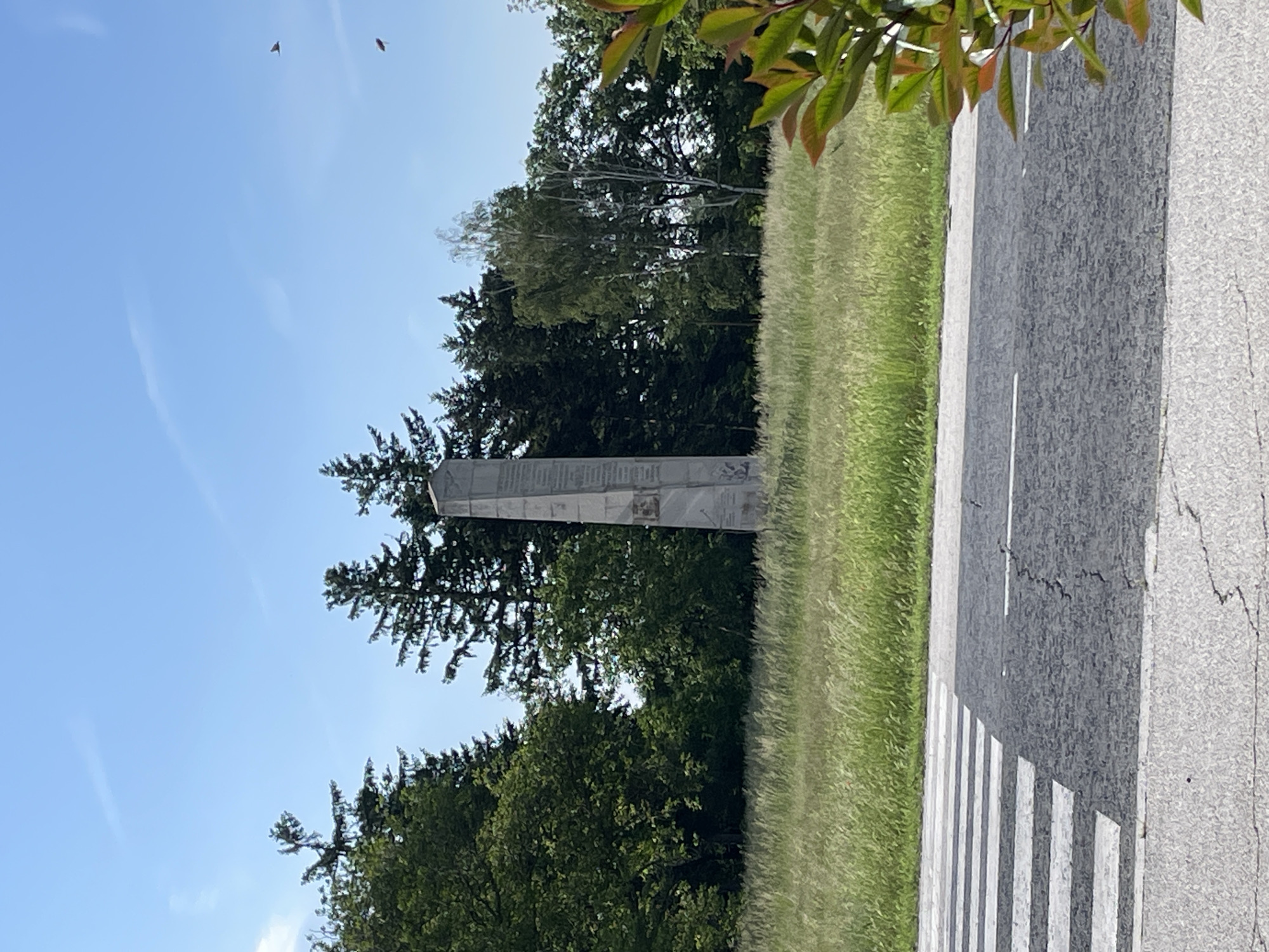 The image shows a tall, gray obelisk situated near a road with a zebra crossing. The obelisk is surrounded by trees and grass. The location appears to be in Sheynovo, Bulgaria, based on the obelisk's design and surrounding environment. There are no people visible. The sky is blue with some light clouds.