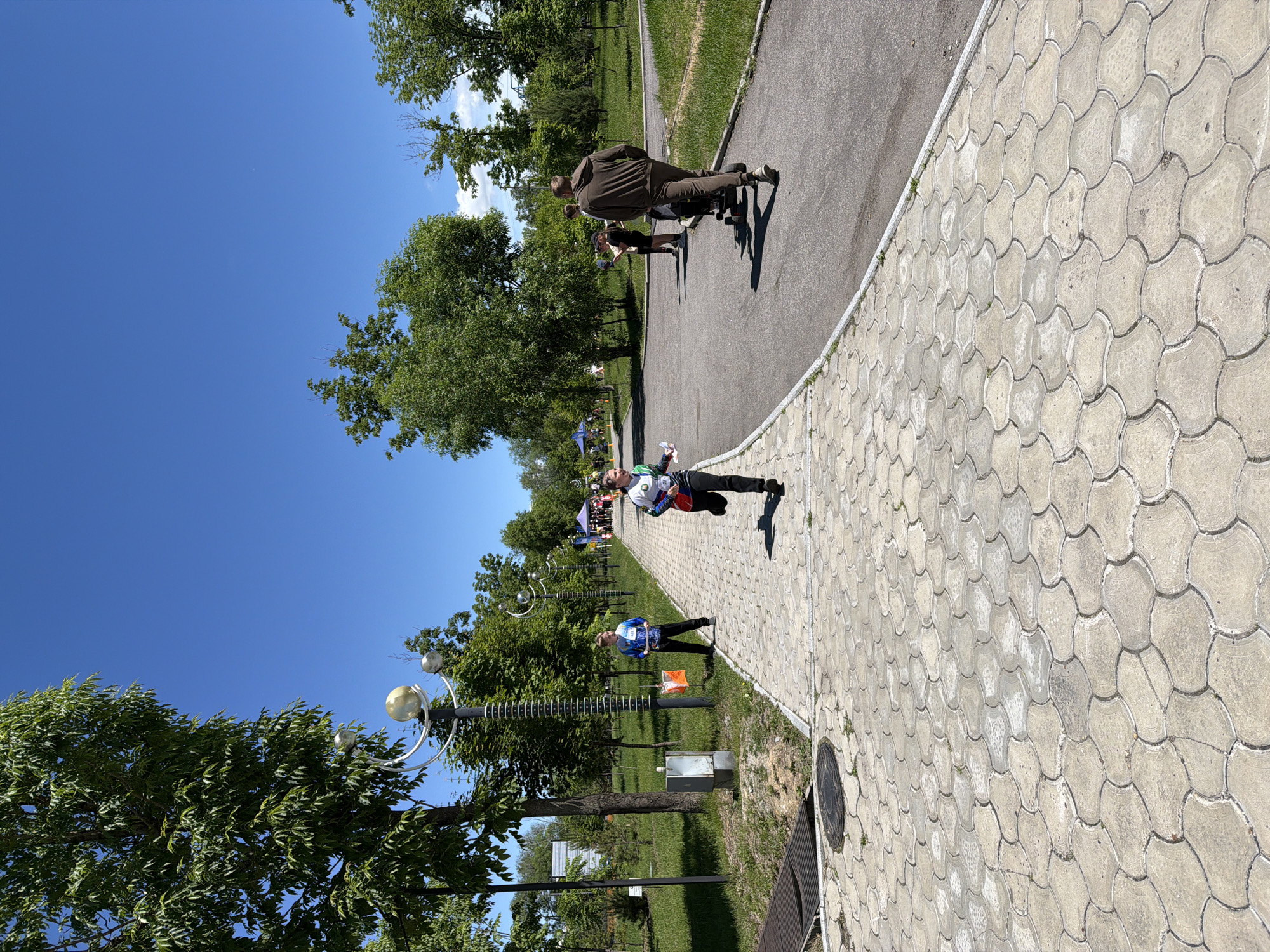 On a sunny day in a park in Khabarovsk, Russia, people are participating in an orienteering event. A young woman in athletic wear is running on a paved pathway, holding a map. Behind her, a man in a blue shirt stands near an orange and white orienteering marker. Other participants are visible in the background, along with trees and park amenities. The pathway is made of interlocking concrete pavers, and a road runs alongside it. The sky is clear and blue.