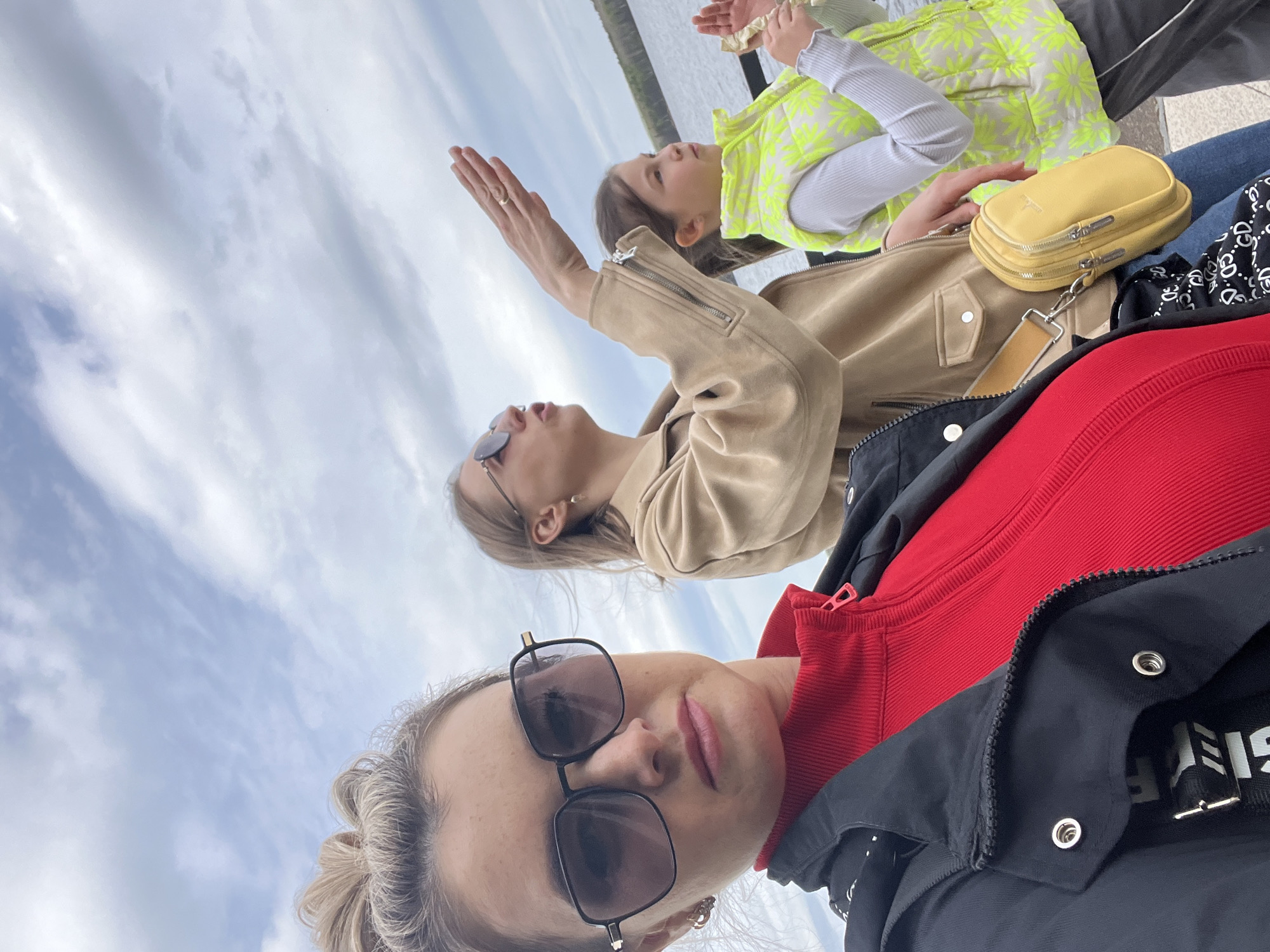 Three individuals are present in the image. The person closest to the camera wears sunglasses, a red top, and a black jacket. The second person wears sunglasses and a tan jacket, with an outstretched hand. A child is in the background wearing a light green and white jacket. A small yellow purse rests near the child. The background shows a body of water and the sky. The location is the city of Chaikovsky, Russia.