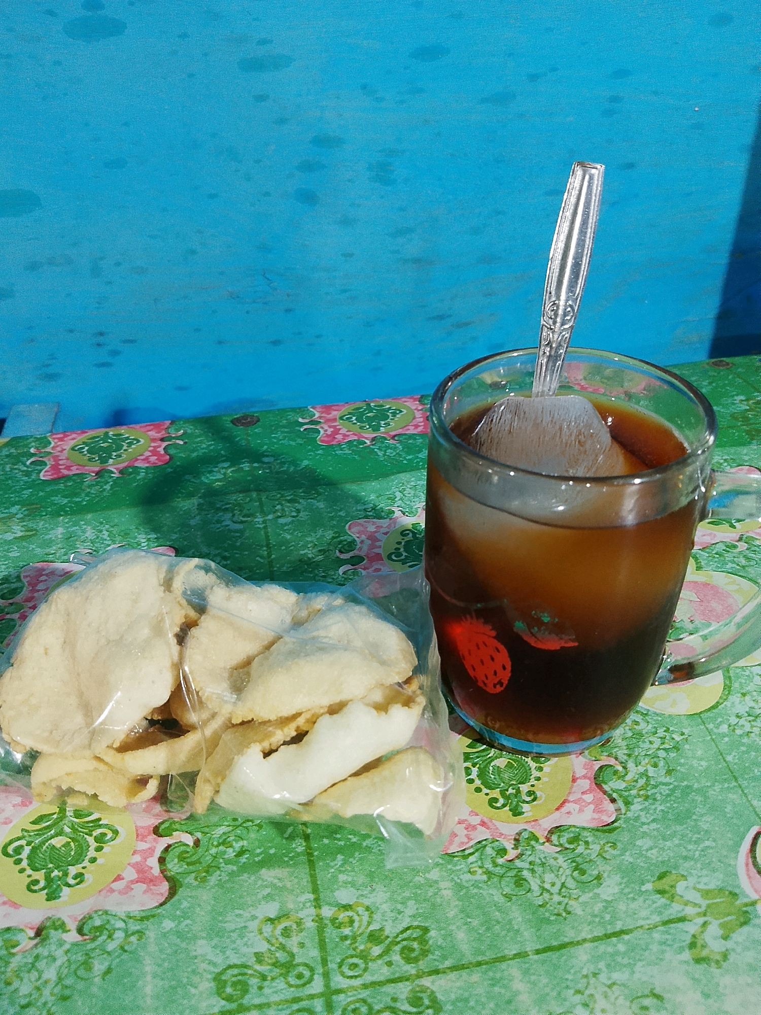 A clear glass mug filled with a dark liquid, possibly iced tea or coffee, rests on a green and pink patterned tablecloth. A large ice cube floats in the beverage, and a silver spoon is inserted into the mug. The glass has a red strawberry design on its side. To the left of the mug is a clear plastic bag containing several light-colored, puffed snacks, resembling crackers or chips. The background is a textured, bright blue surface, possibly a wall or a painted board, with some darker splotches that could be stains or shadows. The lighting suggests an outdoor setting, with shadows cast on the tablecloth. The scene is likely situated outdoors in a casual setting, such as a table on a porch or a street stall, in the city of Pelaihari, Indonesia, given the typical local snacks and beverages. No individuals are visible in the image.