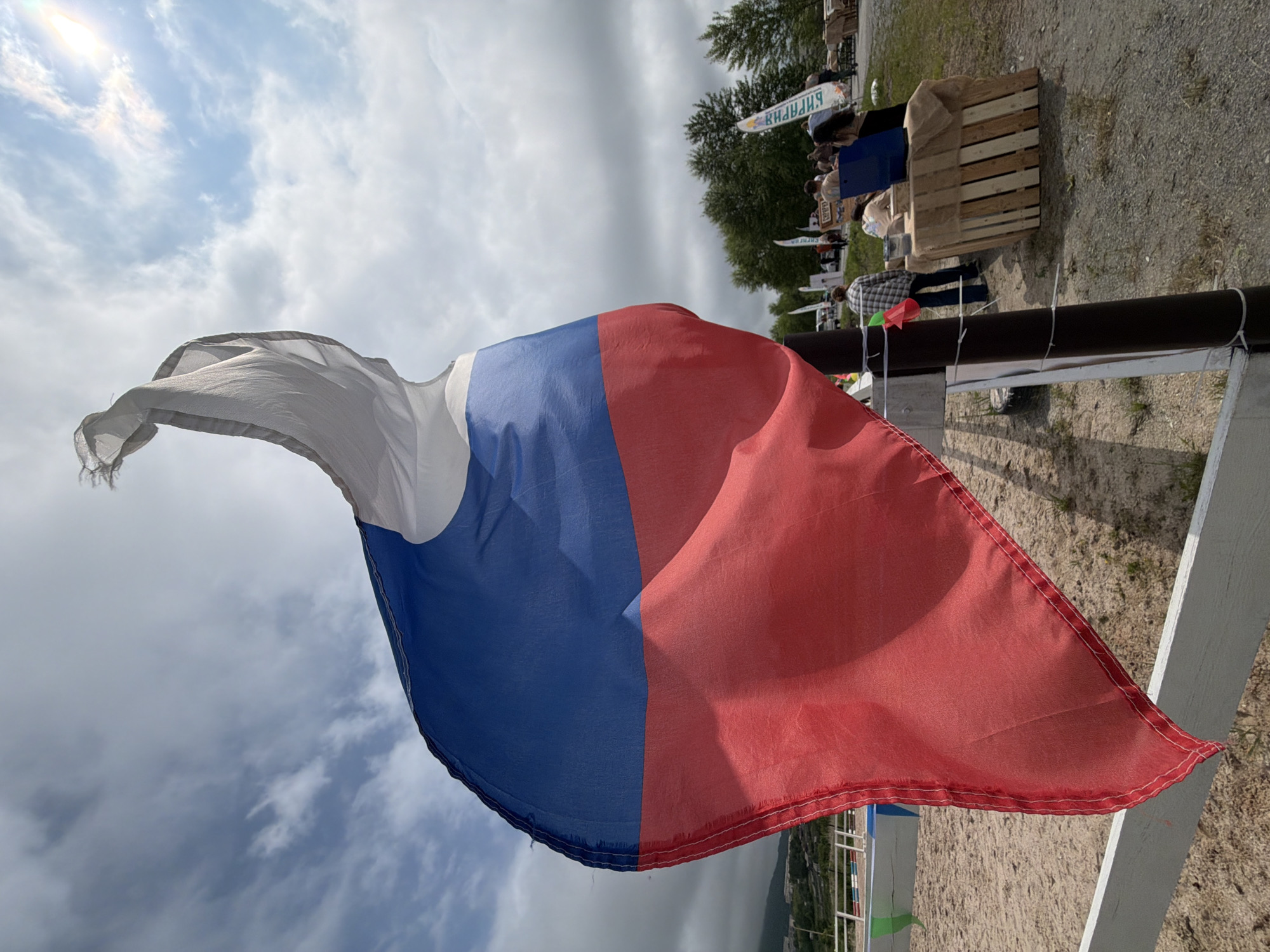 The image shows a Russian flag flying against a cloudy sky. In the background, there are trees, a sand arena, and several people gathered around some stalls or stands. One of the flags in the background has Cyrillic writing. The overall scene suggests an outdoor event or fair in a rural or semi-rural setting.