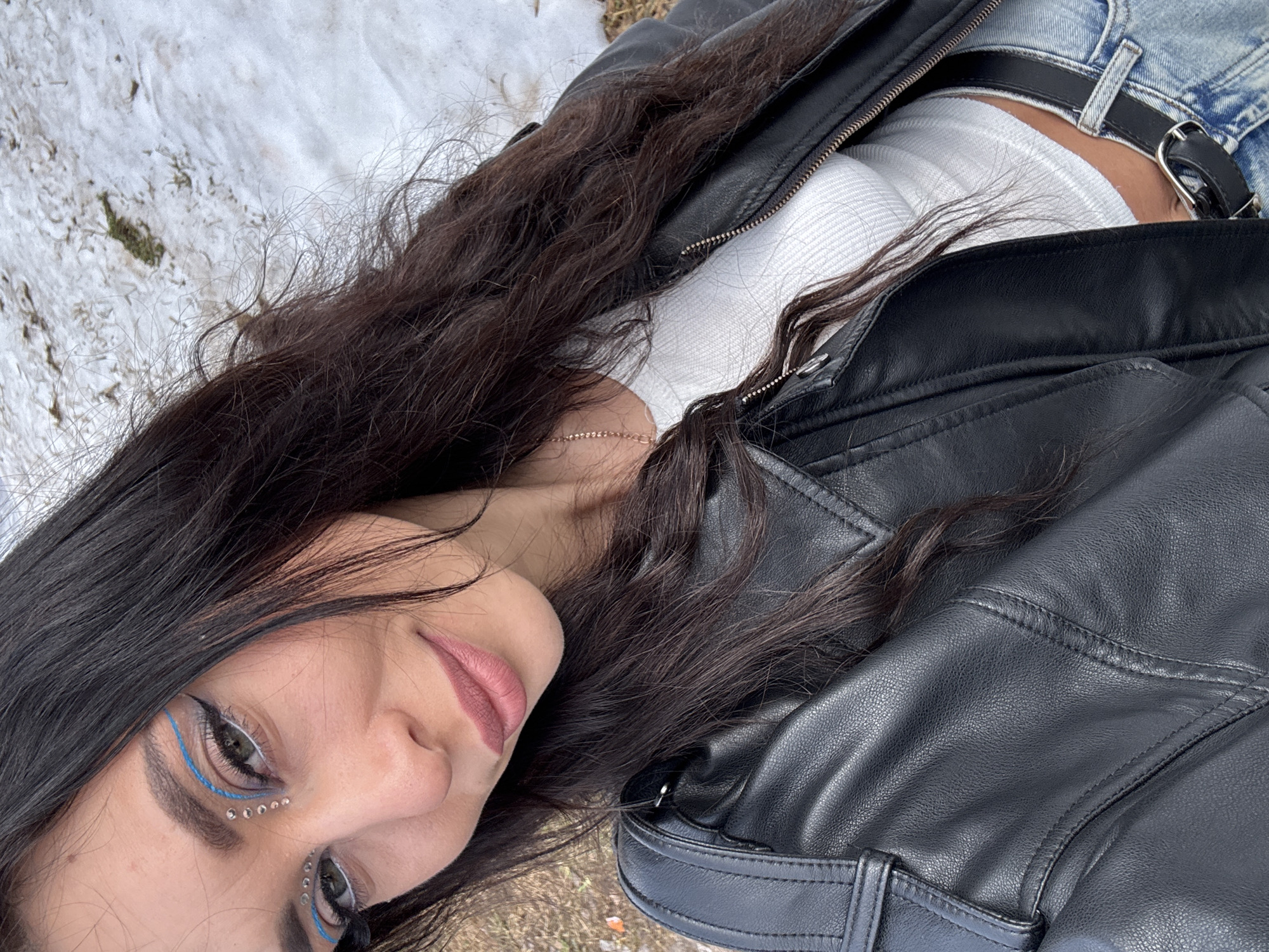 A woman lies on her side outdoors in what appears to be a snowy, grassy area. She has long, dark hair and is wearing blue eyeliner with decorative jewels under her eyes. She is also wearing a white top, blue jeans with a black belt, and a black leather jacket. There is snow visible in the background. The location is not Фоки, Russia.