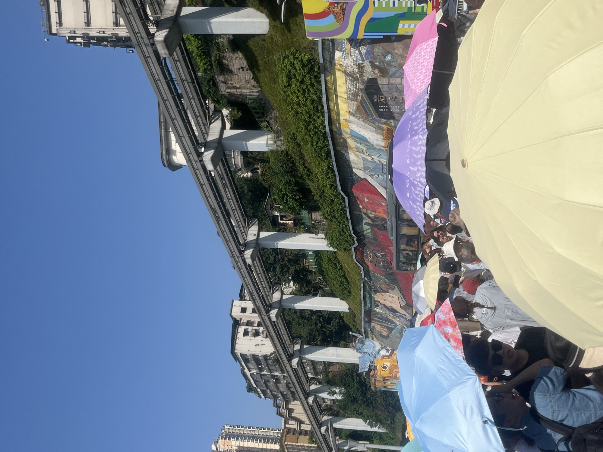 The image presents a street scene in Yuzhong District, China. A monorail track runs across the upper portion of the frame, supported by white pillars. Buildings are visible on either side of the track. A long mural extends along a retaining wall.
A crowd of people is gathered in the foreground, many holding umbrellas, including colors such as light yellow, purple, pink, and blue. Some people are wearing sunglasses. Details of individual interactions are obscured by the density of the crowd and the umbrellas.