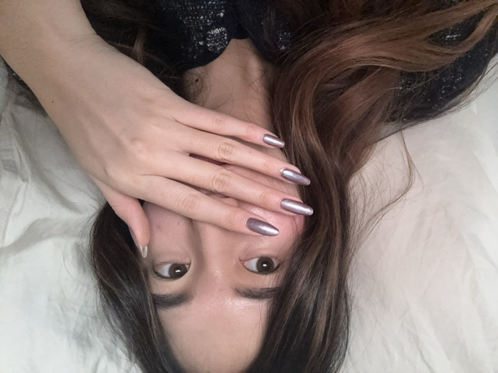 The image displays a close-up portrait of a female individual situated in an indoor environment. The subject has light-to-medium skin, dark brown hair with visible reddish-brown highlights, and dark brown eyes directed slightly towards the upper left of the frame. Her left hand is positioned over the lower part of her face, specifically covering her mouth and chin. The nails on her left hand are long, almond-shaped, and painted with a metallic, shimmery purplish-silver polish. She is wearing a dark-colored, textured garment, possibly knit. The background consists of light-colored, crumpled fabric, consistent with bedsheets or bedding material. The lighting is soft and even. No visual information within the image identifies the geographical location as Paris, France, or any other specific city.