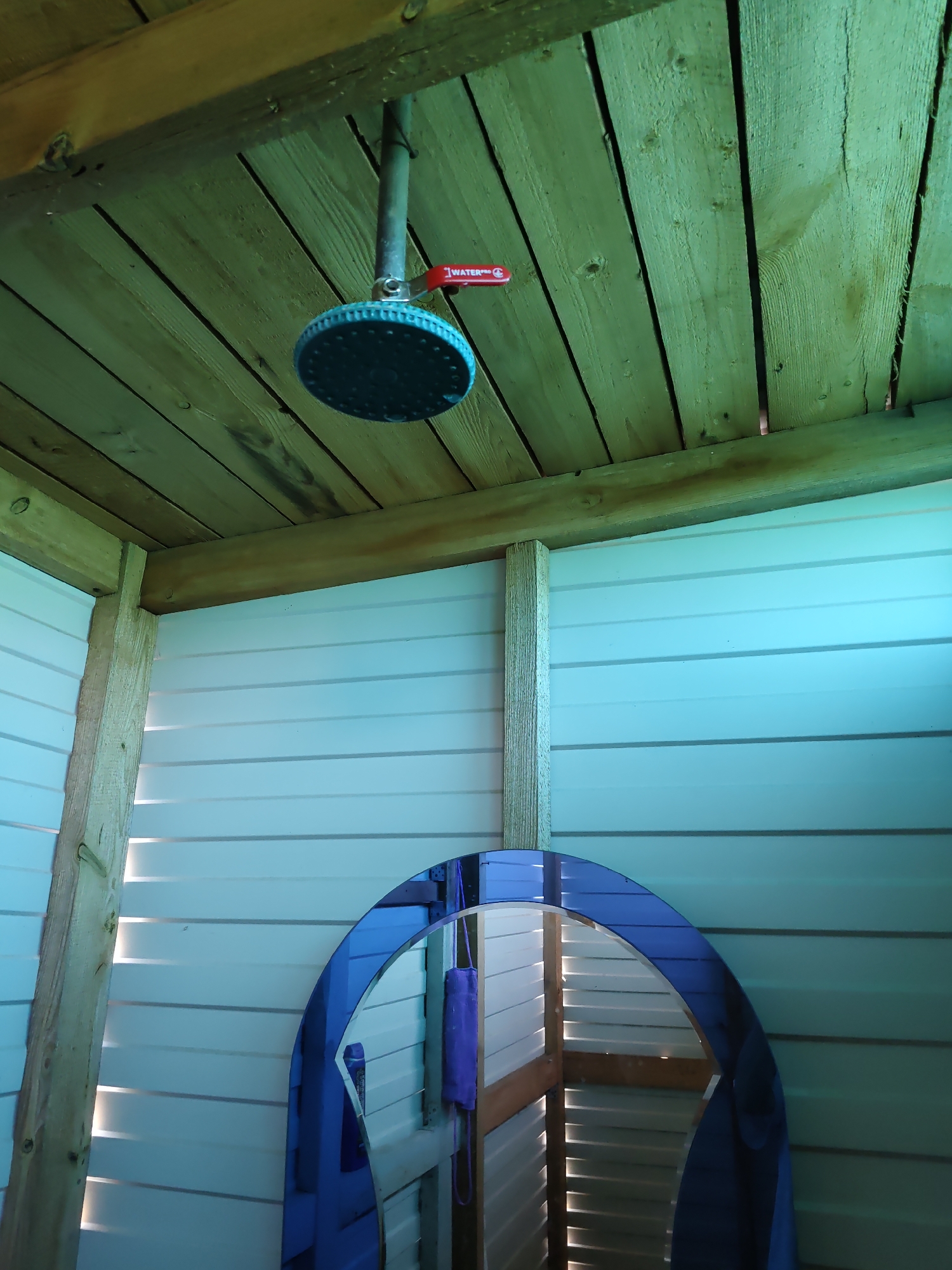 This is an interior shot of an outdoor shower stall. The ceiling is made of wood planks, and the walls are constructed of white horizontal paneling. A round showerhead with a red valve is visible hanging from the ceiling. In the foreground, a large oval mirror with a blue frame is leaning against the wall, reflecting the interior of the stall, including a purple cloth hanging on the wall. The overall lighting suggests it is daytime. The image appears to be taken in Nizhnedonskoy, Russia, but there are no specific identifying landmarks to confirm this location within the image itself.