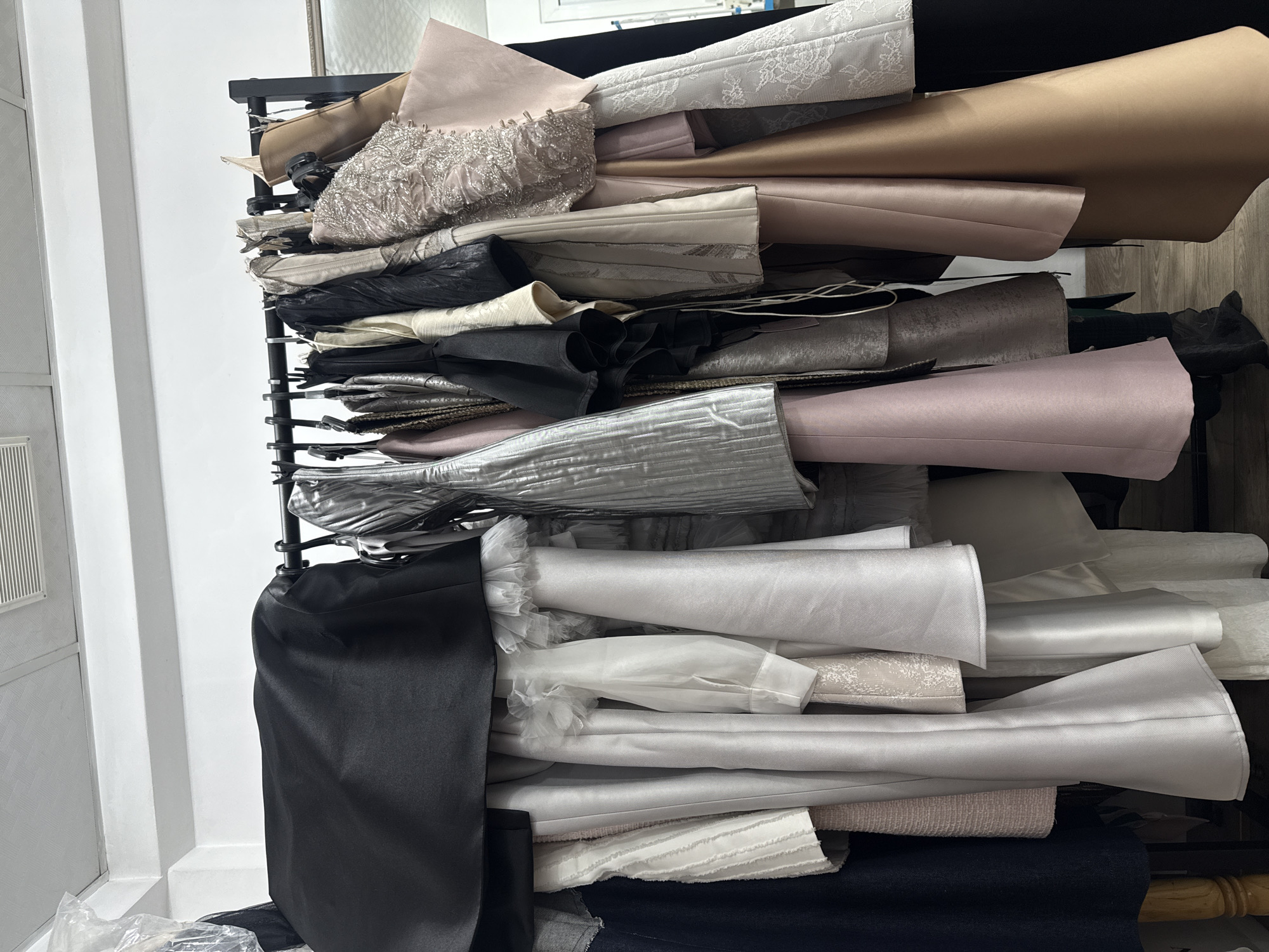 The image shows a metal clothing rack heavily laden with fabric rolls of various colors, textures, and materials. Predominantly, the fabrics include shades of white, silver, pink, brown, and black. Some fabrics appear to be satin, while others have embellishments like lace or beaded details. The rack is positioned indoors, near a white wall with a paneled design. The lighting is diffused, suggesting an interior setting. There is no visible information to confirm the location in the city of Yerevan, Armenia.