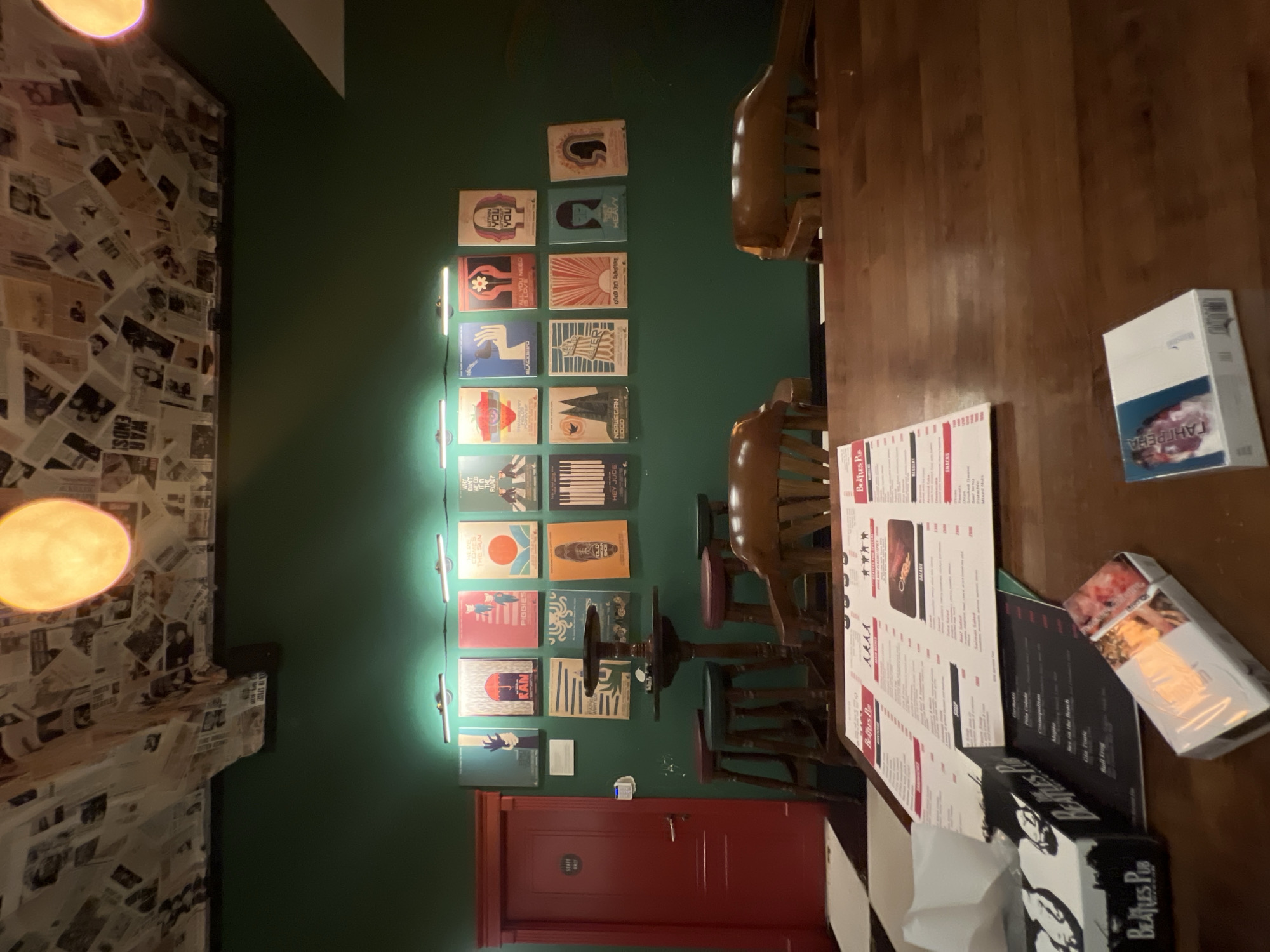 The image depicts the interior of a dimly lit pub with a dark green wall adorned with framed posters. On the ceiling, numerous newspaper clippings are pasted, and a circular, glowing light fixture is visible. A wooden table occupies the foreground, laden with a menu, a box of tissues with "Beatles Pub" branding, and a pack of cigarettes. Several wooden chairs are arranged around the table, and a red door with a "Staff Only" sign is present on the left side of the image. A checkered floor pattern is visible in the lower left corner. The overall ambiance suggests a casual, retro-themed establishment. The location is identified as The Beatles Pub in Yerevan, Armenia.