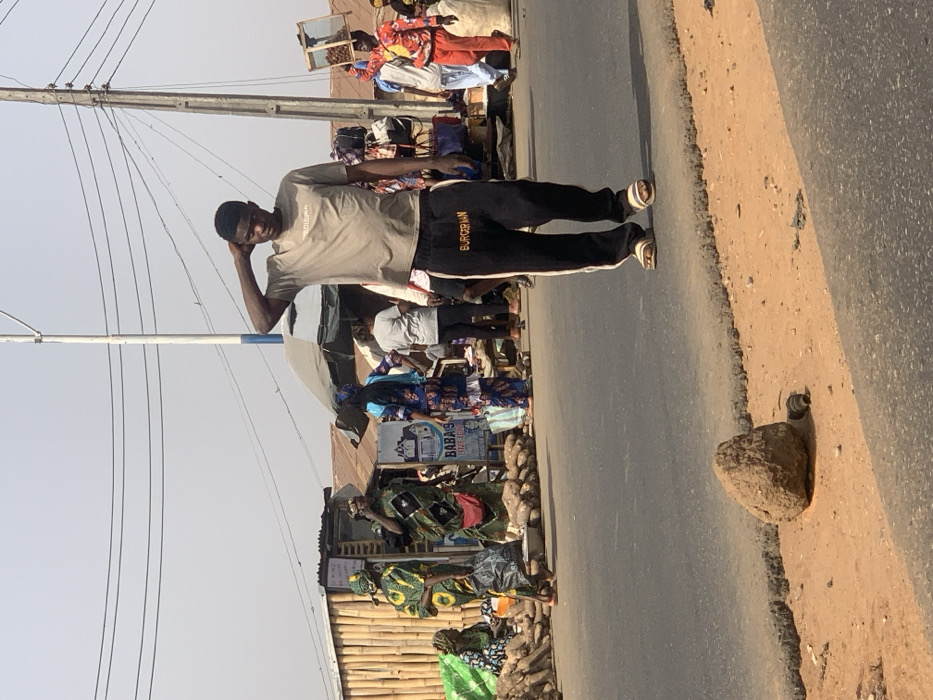 The image, originally rotated 90 degrees clockwise, captures a bright daytime street scene in Amoyo, Nigeria, under a clear sky. A young man stands prominently on the edge of an asphalt road, part of his stance on the dirt shoulder. He wears a light grey t-shirt, dark sweatpants with the word "BURGUIGNON" printed vertically on the left leg, and sandals. His right hand is resting on the back of his head or neck, and he appears to be looking towards the left side of the frame.
To his left, a bustling market or street vendor area is visible. Numerous people, predominantly women in colorful, patterned clothing, are engaged in market activities around various stalls. Goods, including what appear to be large piles of yams, are displayed on the ground. Simple structures, possibly with corrugated iron roofs, serve as market stalls. One sign is partially visible, showing "BABA'S" and "STORE", while another sign has some indiscernible text that includes "HOUSE". In the background, a vendor is seen carrying a wooden tray on their head.
To the man's right, a tall utility pole with multiple power lines rises against the sky. The road features a distinct dirt shoulder where a large rock and a discarded bottle or can are present. The strong sunlight casts clear shadows, indicating a mid-day to late afternoon setting. The overall scene depicts a vibrant street market environment typical of the region.
