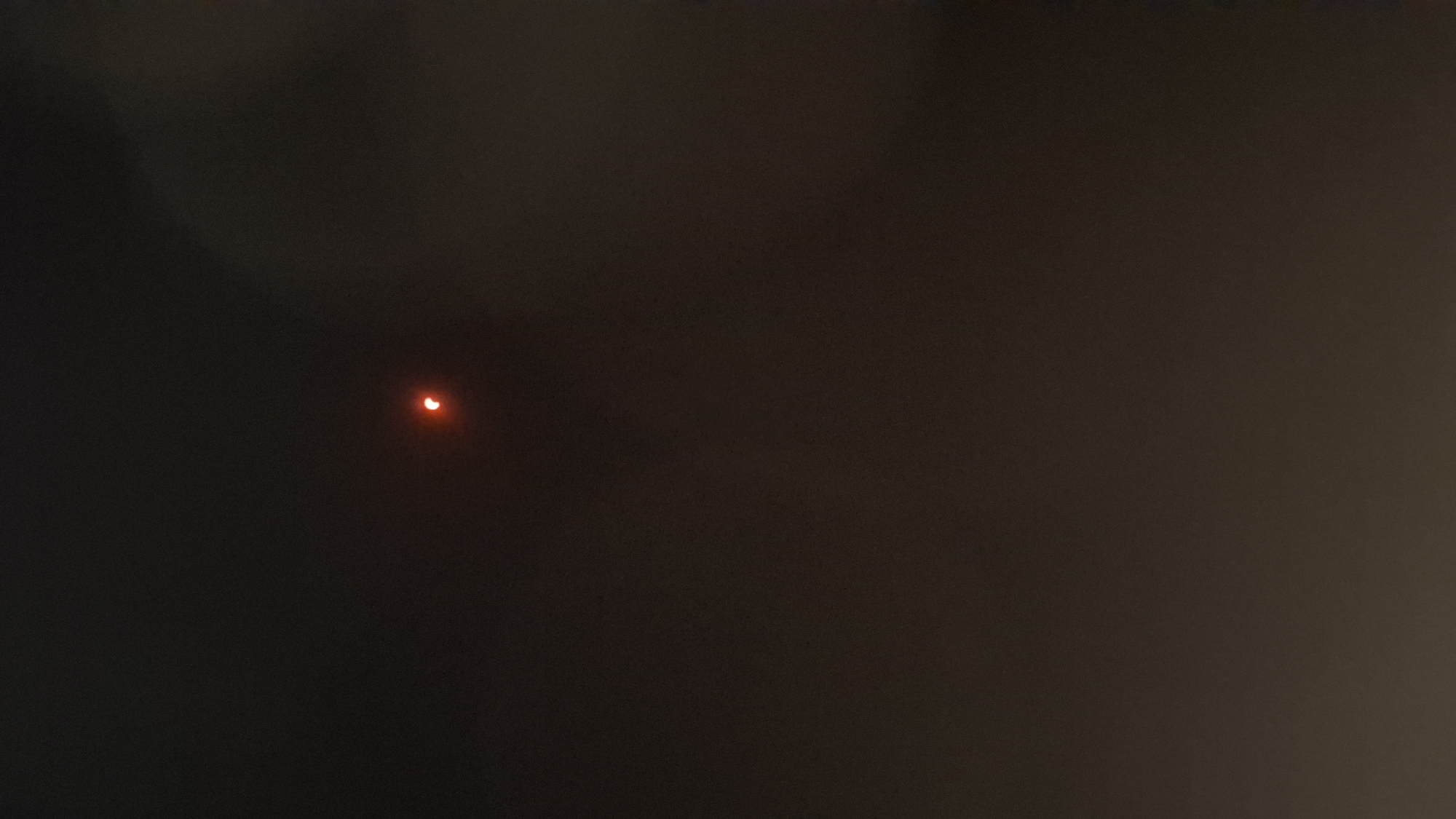 The image shows a partial solar eclipse. A bright, crescent-shaped sliver of the sun is visible against a dark sky, likely at dusk or night. The sliver appears to be orange or red, possibly due to atmospheric conditions or filtering. The rest of the sky is dark and appears slightly hazy or cloudy. The scene is devoid of any discernible human presence, urban structures, or specific landmarks that would indicate the city of Híjar, Spain.