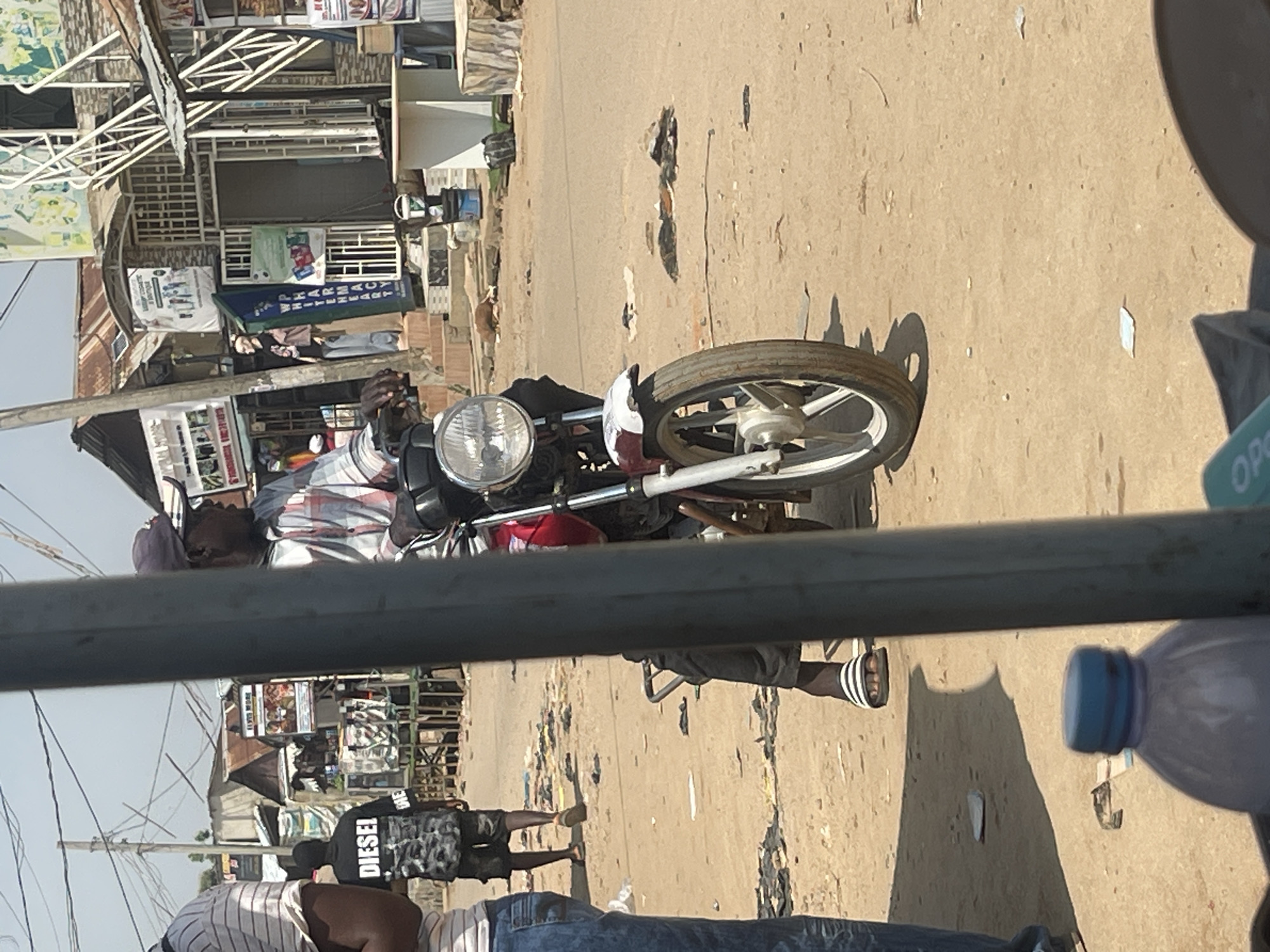 A medium shot captures a street scene in Abuja, Nigeria, with a man seated on a red and white motorcycle. He is wearing a red, white, and blue plaid shirt, a dark baseball cap, and glasses, with a portion of his face visible as he looks to his right. His right leg, clad in black pants and a striped sandal, is extended forward, resting on the ground near the motorcycle's kickstand. The motorcycle, a red and white model with a prominent front headlight and spoked wheel, is positioned in the foreground. Behind the man and motorcycle, a dusty, unpaved road is visible, littered with scattered debris. To the left of the motorcycle, a person wearing blue jeans and a striped t-shirt stands with their back partially to the camera. Further in the background, several small shops and structures line the street, with some displaying signage and advertisements. Power lines crisscross overhead, and the overall environment appears to be a bustling, informal commercial area under a clear sky.